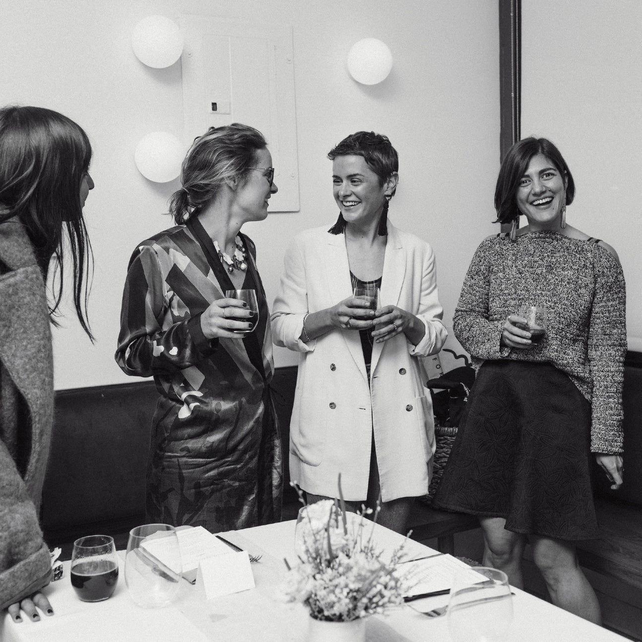 Black and white photo of Lizzie and Kathryn with friends, holding wine glasses behind a table