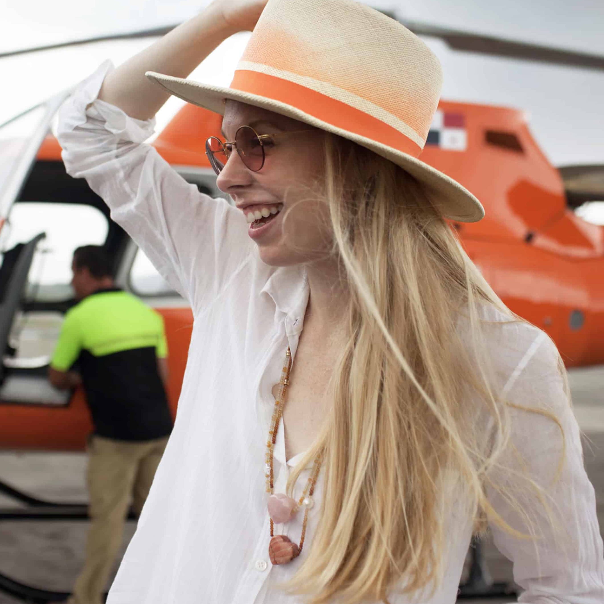 Gigi Burris stands in front of helicopter holding her straw hat. She wars white button-down with necklaces.