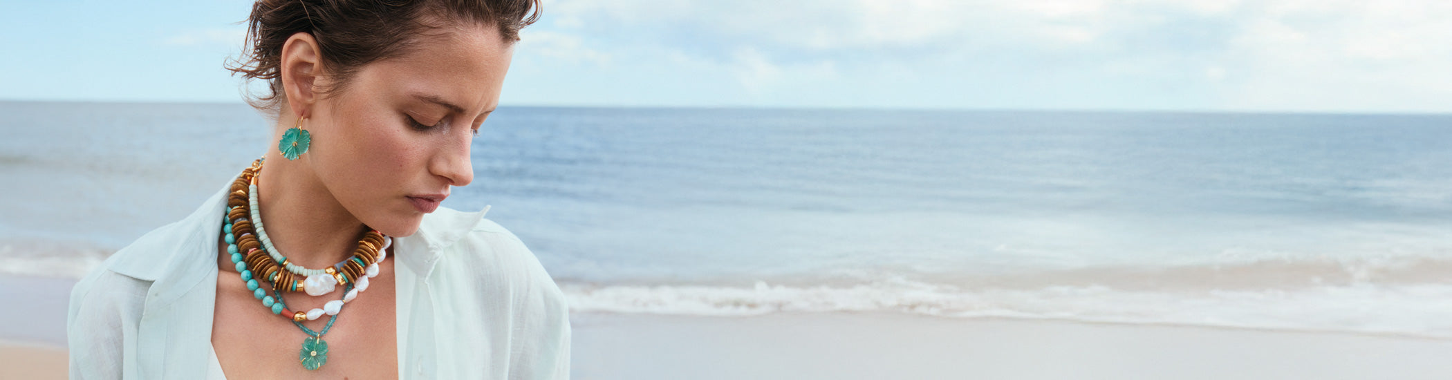 Model on beach wears aqua button-down with stack of bead necklaces and New Bloom Earrings in Capri