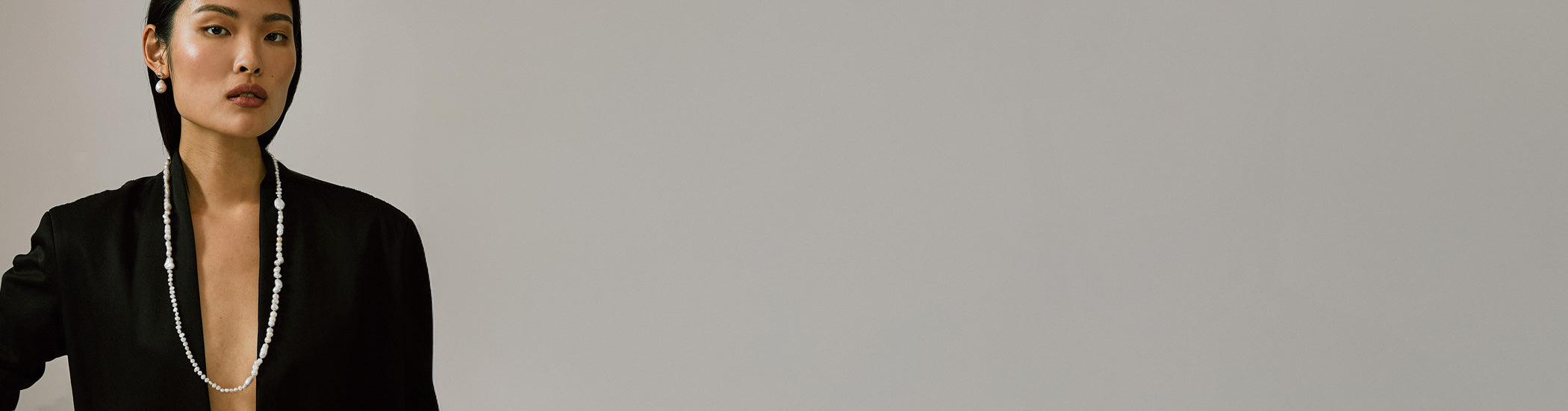 Model on grey backdrop wears black blazer with long pearl necklace