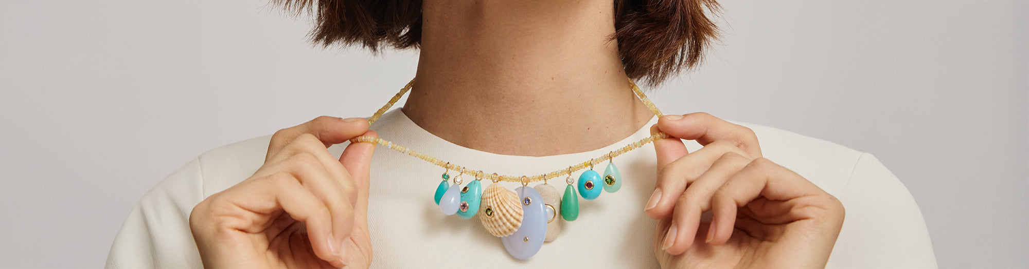 Model on grey backdrop wears cream top with beaded opal necklace laden with semiprecious charms.