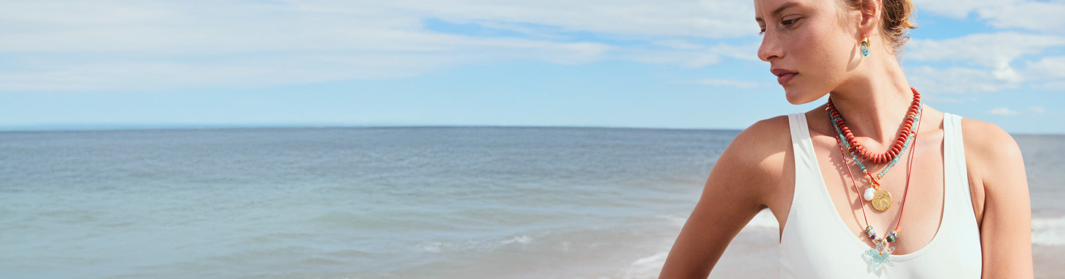 Model on beach wears white tank top with assorted blue and red necklaces
