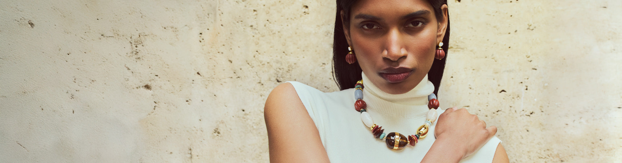 Model on stone backdrop wears cream sleeveless top with Kona Collar and Pepo Earrings