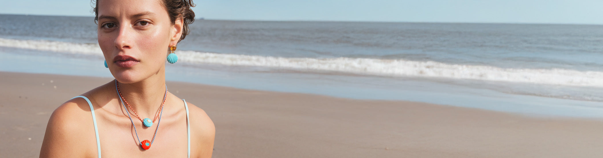 Model on beach wears Kai Earrings and Necklaces in Red Hot and Ultramarine