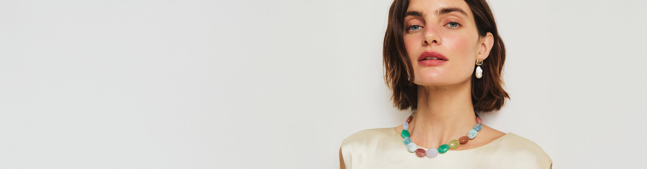 Model on grey backdrop wears cream silk top with Mixed Gemstone Necklace and Juno Earrings.
