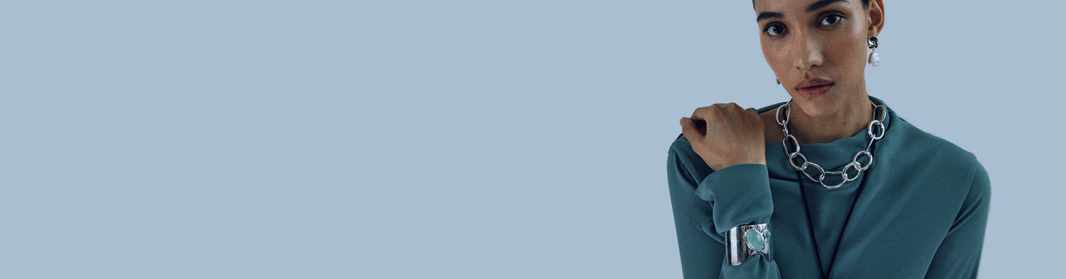 Model on blue backdrop wears teal top with Porto Chain, Coin Reflection Earrings and Organic Cuff in Silver