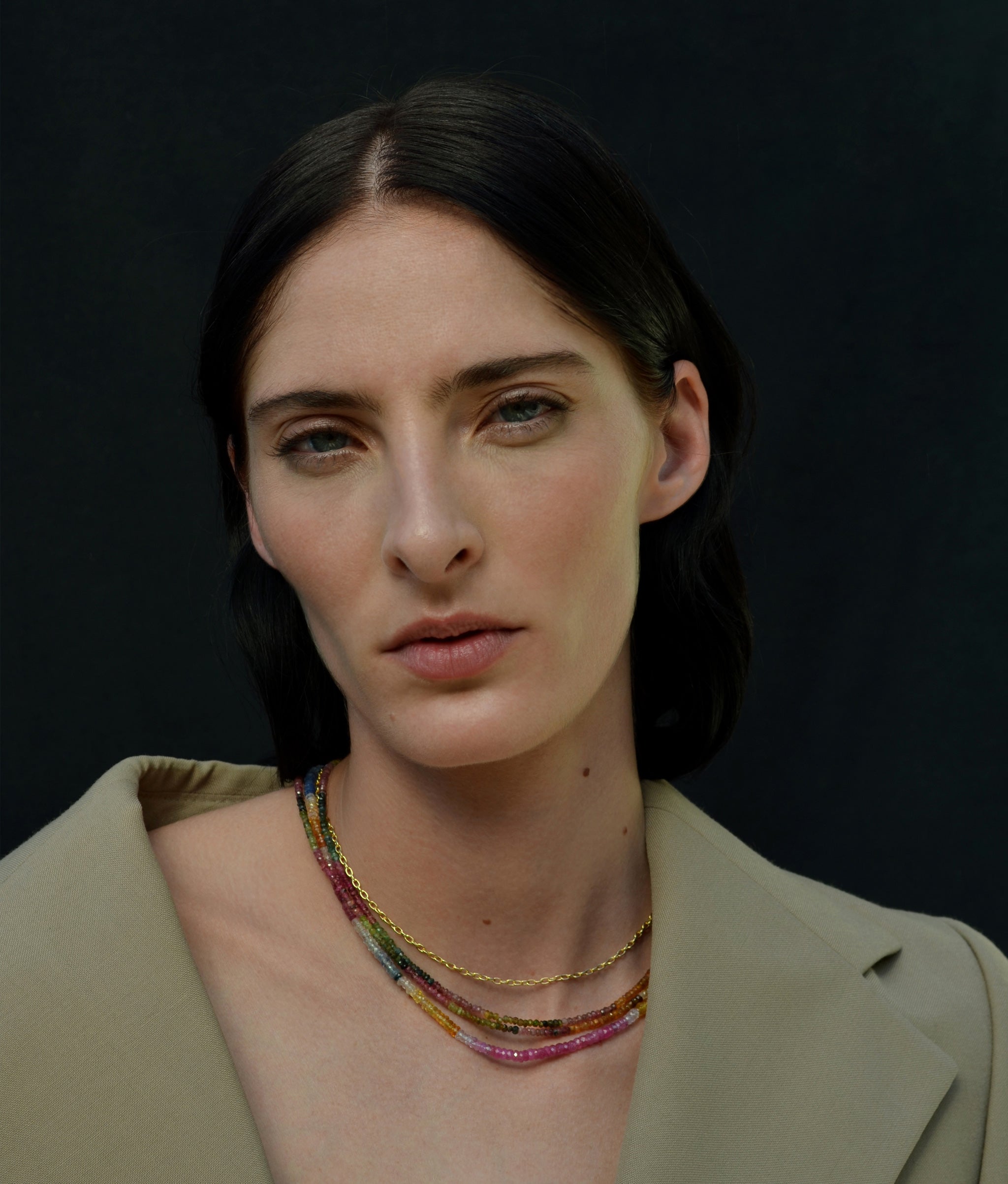 Model in olive jacket wears various beaded and gold necklaces.