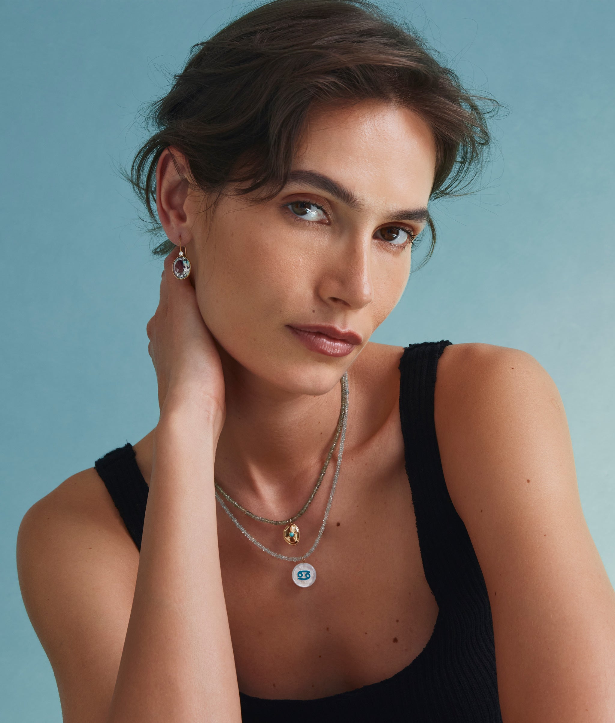 Model on blue backdrop wears black tank with tiny beaded necklaces and Birthstone charm in Turquoise