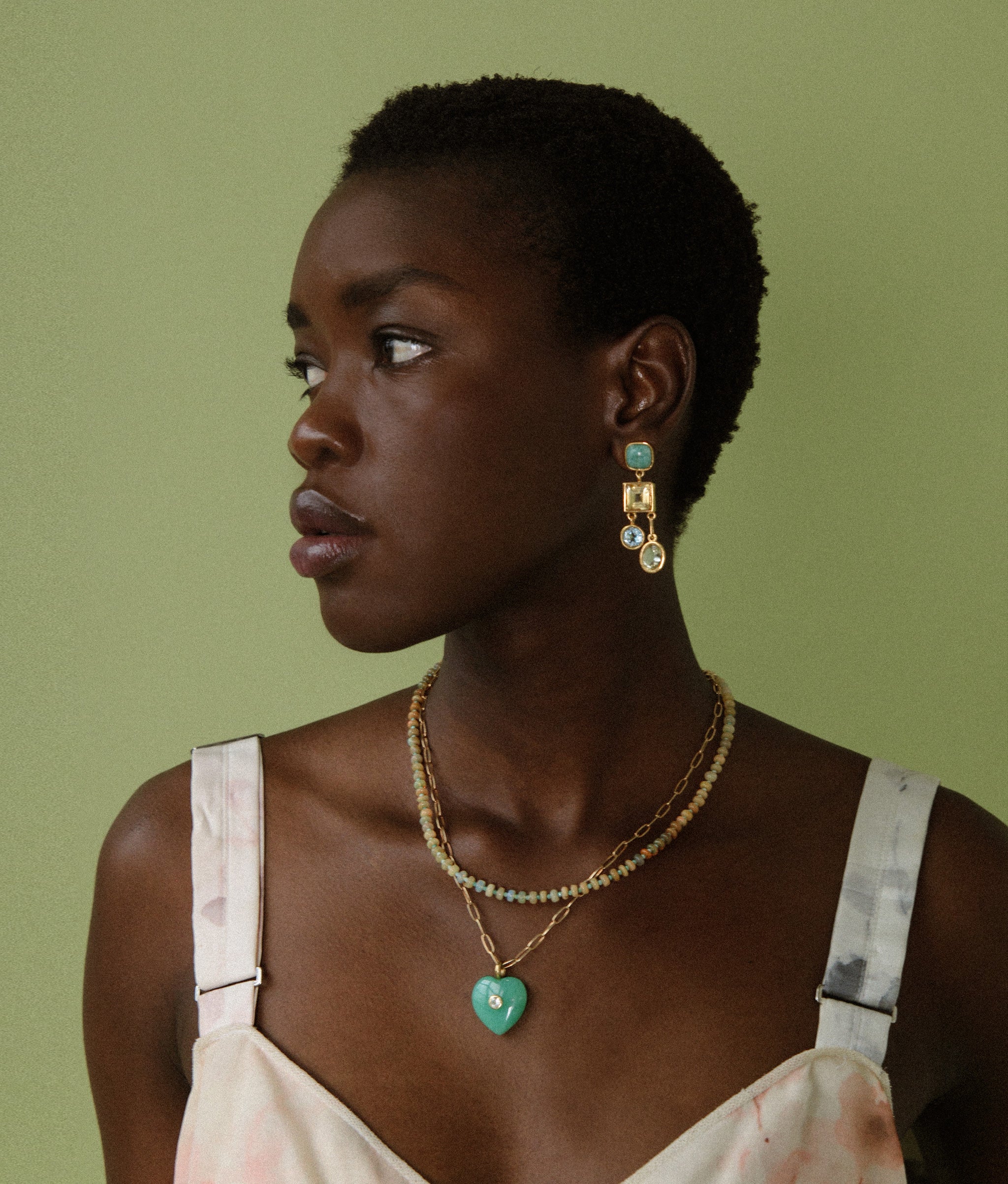 Model in profile on green backdrop wears floral dress with Anemone Earrings and Lamai Heart Necklace.