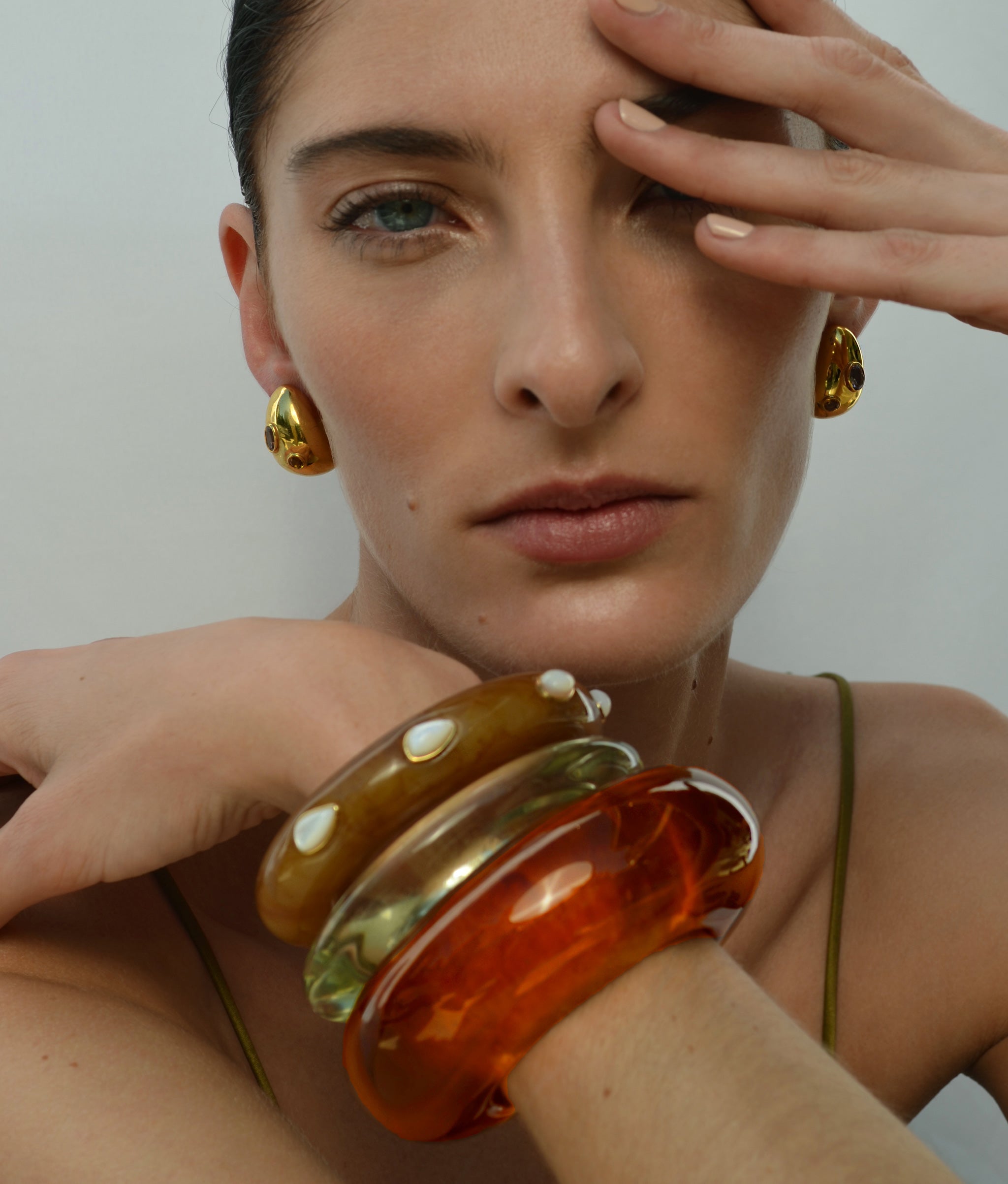Model on grey backdrop wears stack of resin bracelets with Mini Arp Earrings in Studded Gold.