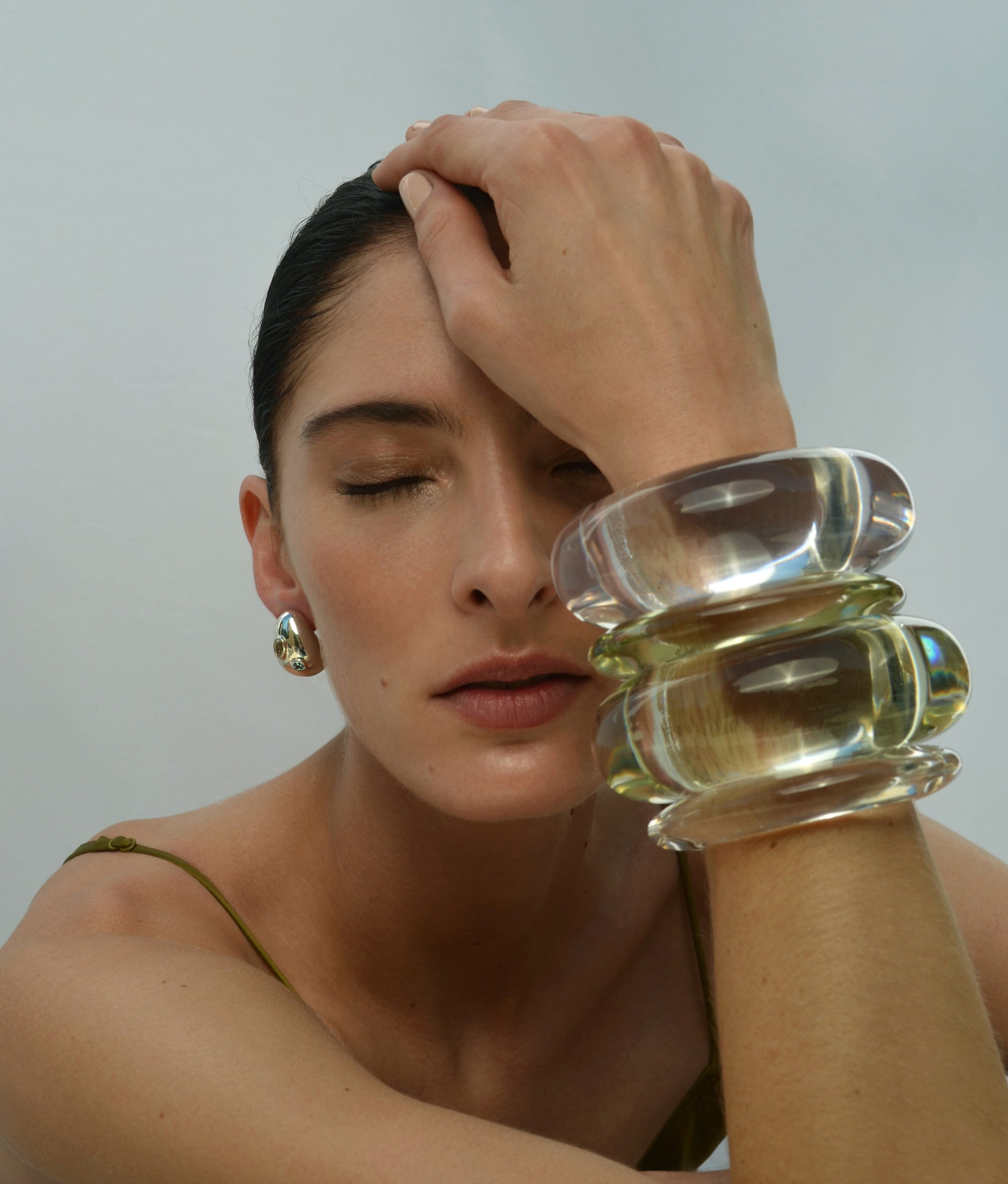 Model on grey backdrop puts hand to head with stack of resin bracelets, wears Mini Arp Earrings