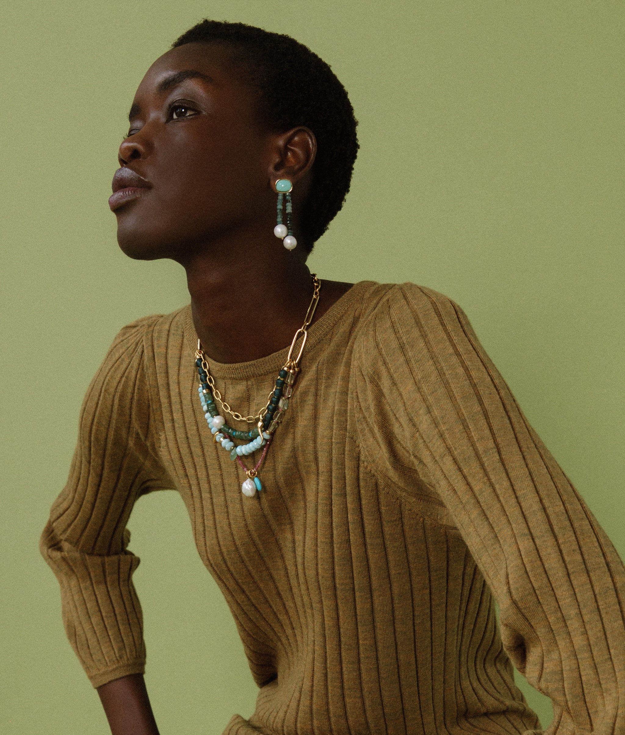 Model on green backdrop wears mustard knit top with Alvar Earrings and Vizcaya Necklace.