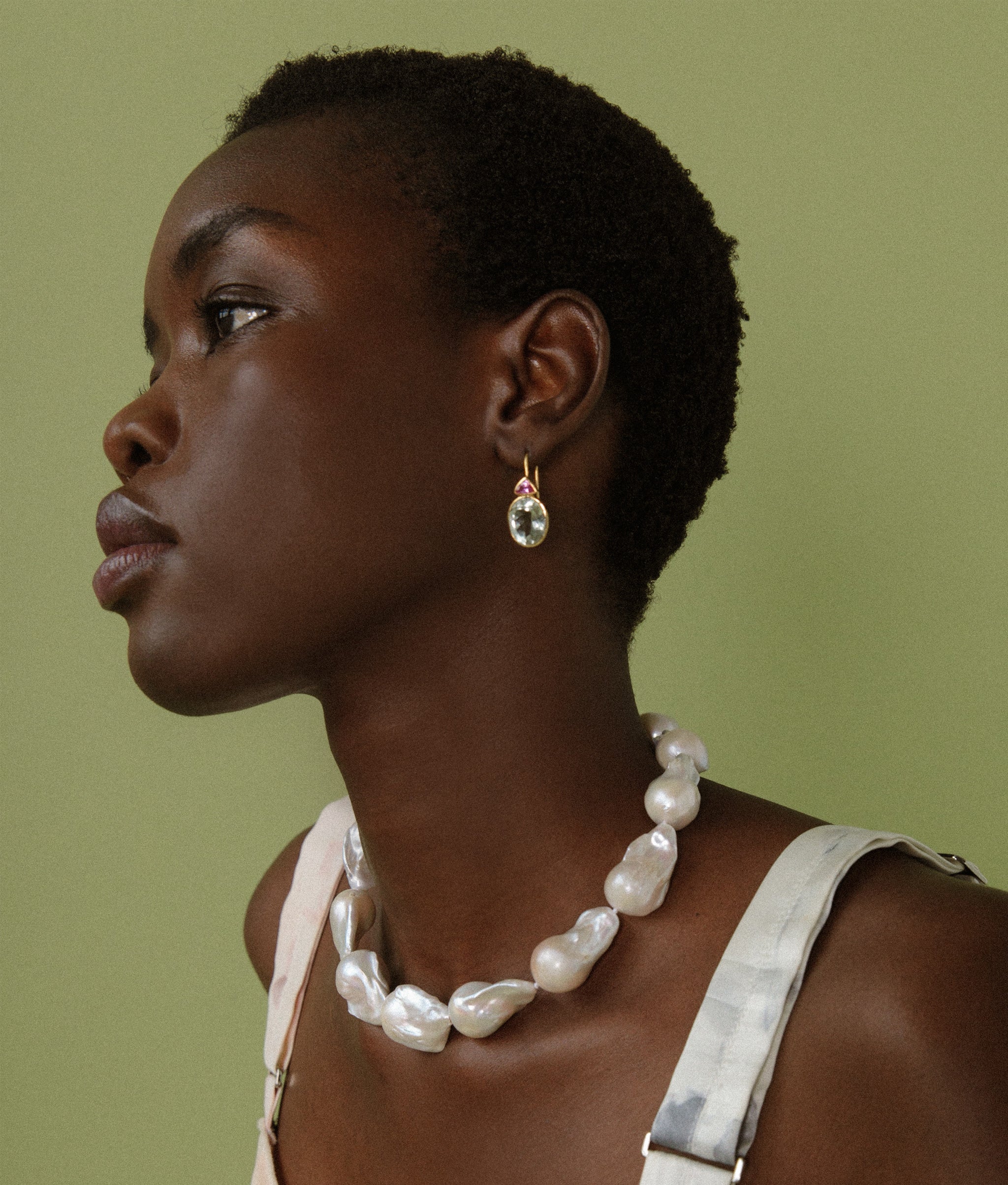 Model on green backdrop wears Extra Large White Baroque Pearl & 14k Gold Necklace with fine earrings and white top.