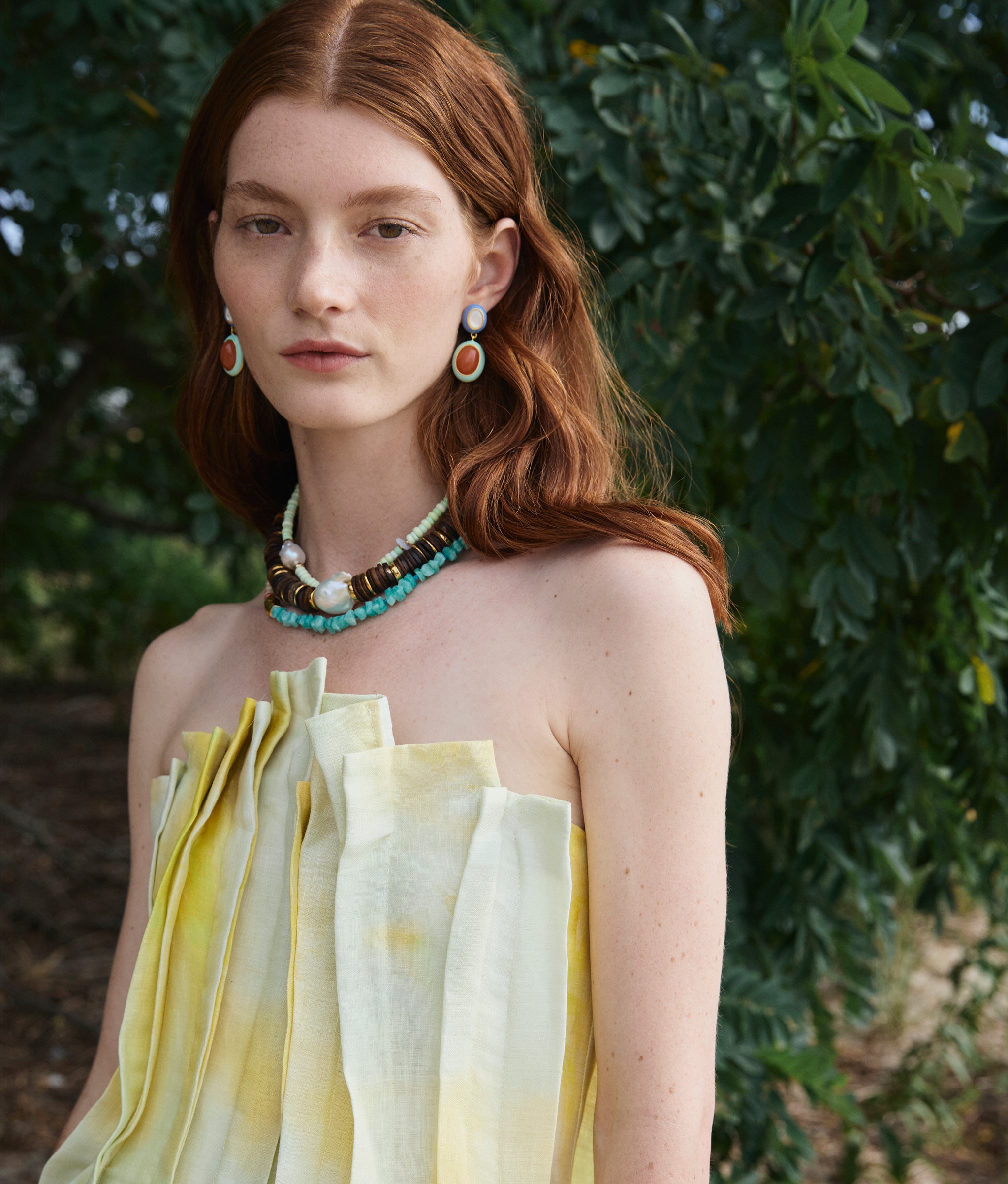Model in strapless top wears Papaya Earrings in Peach, Bilbao Collar II, Cabana Necklace in Honeydew in front of a tree.