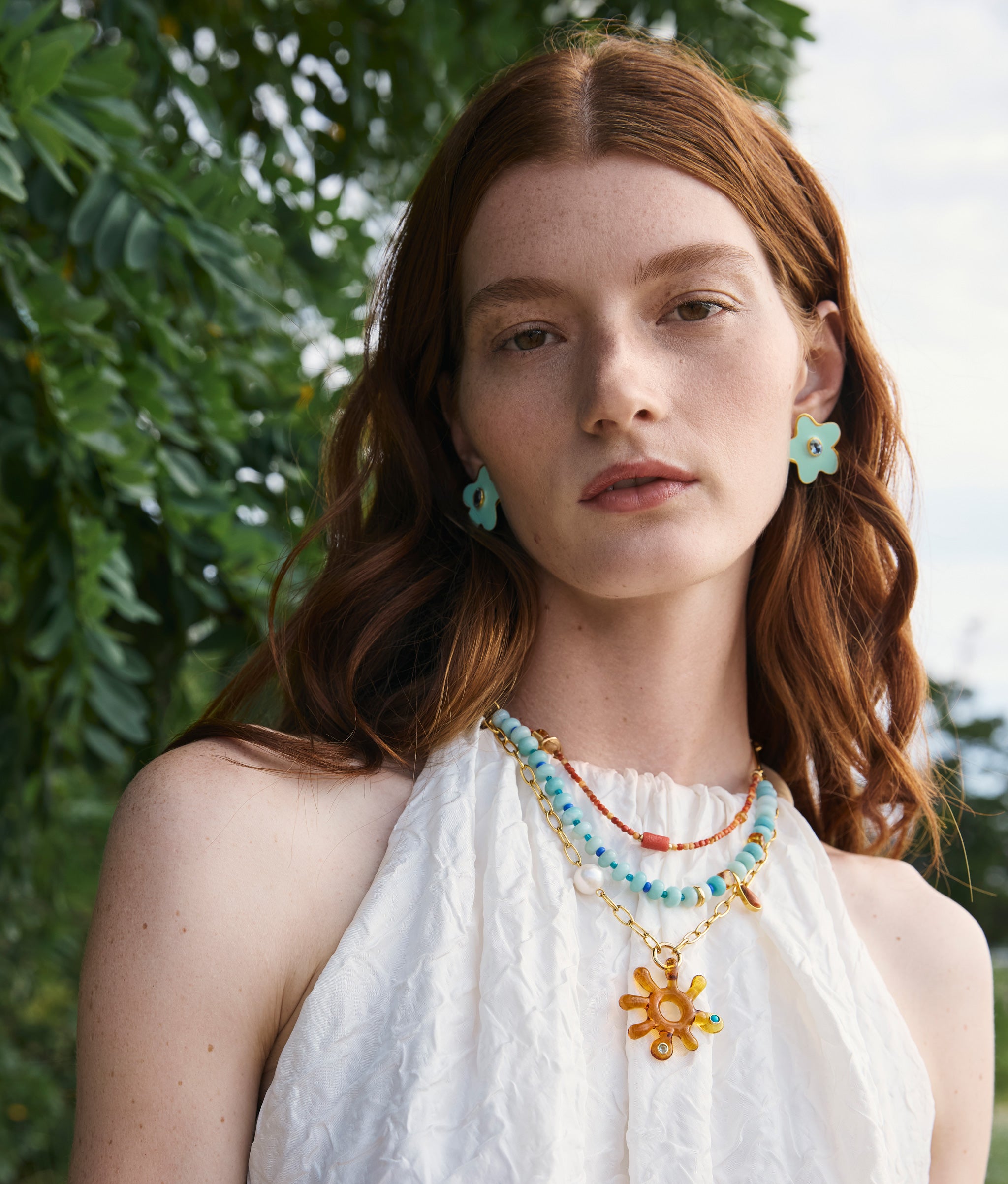 Model wears white blouse, Sunburst Necklace and Camilo stud earrings, atop a leafy background