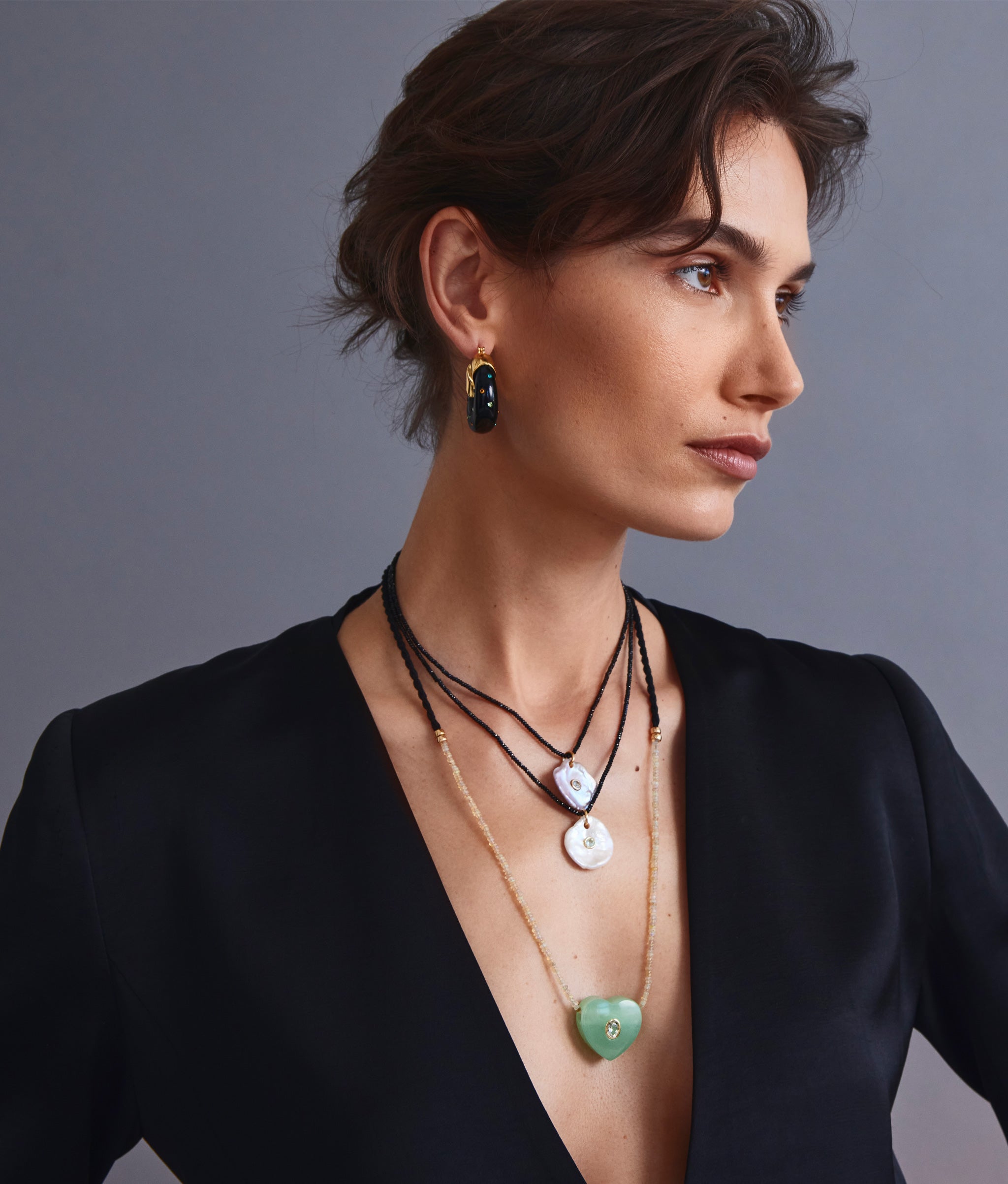 Model on grey backdrop wears black blazer with Gemini Necklace in Mint, two Meret Necklaces, and Organic Hoops.