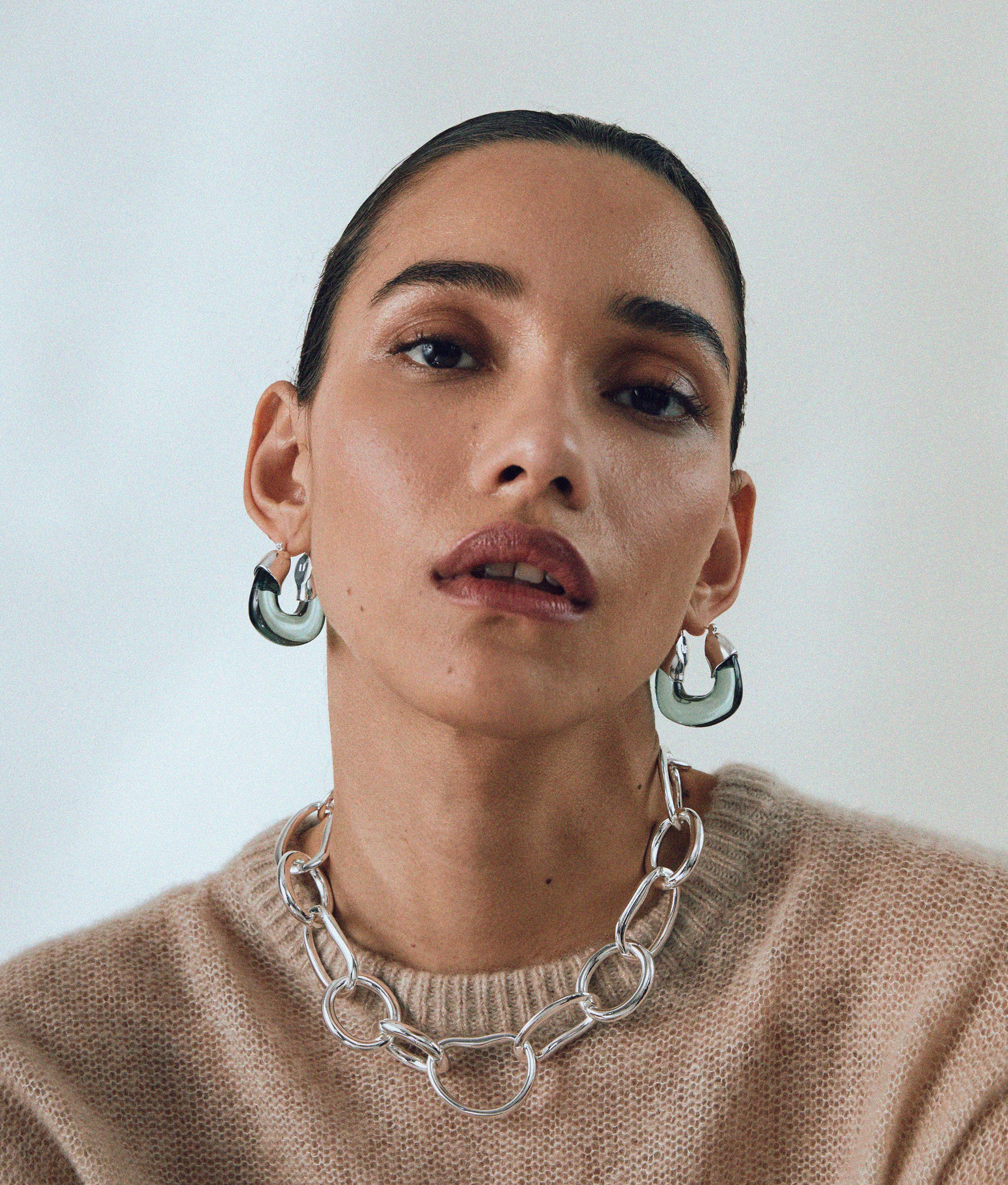 Model in tan sweater wears Porto Chain in Silver and Organic Hoops in Forest & Silver.