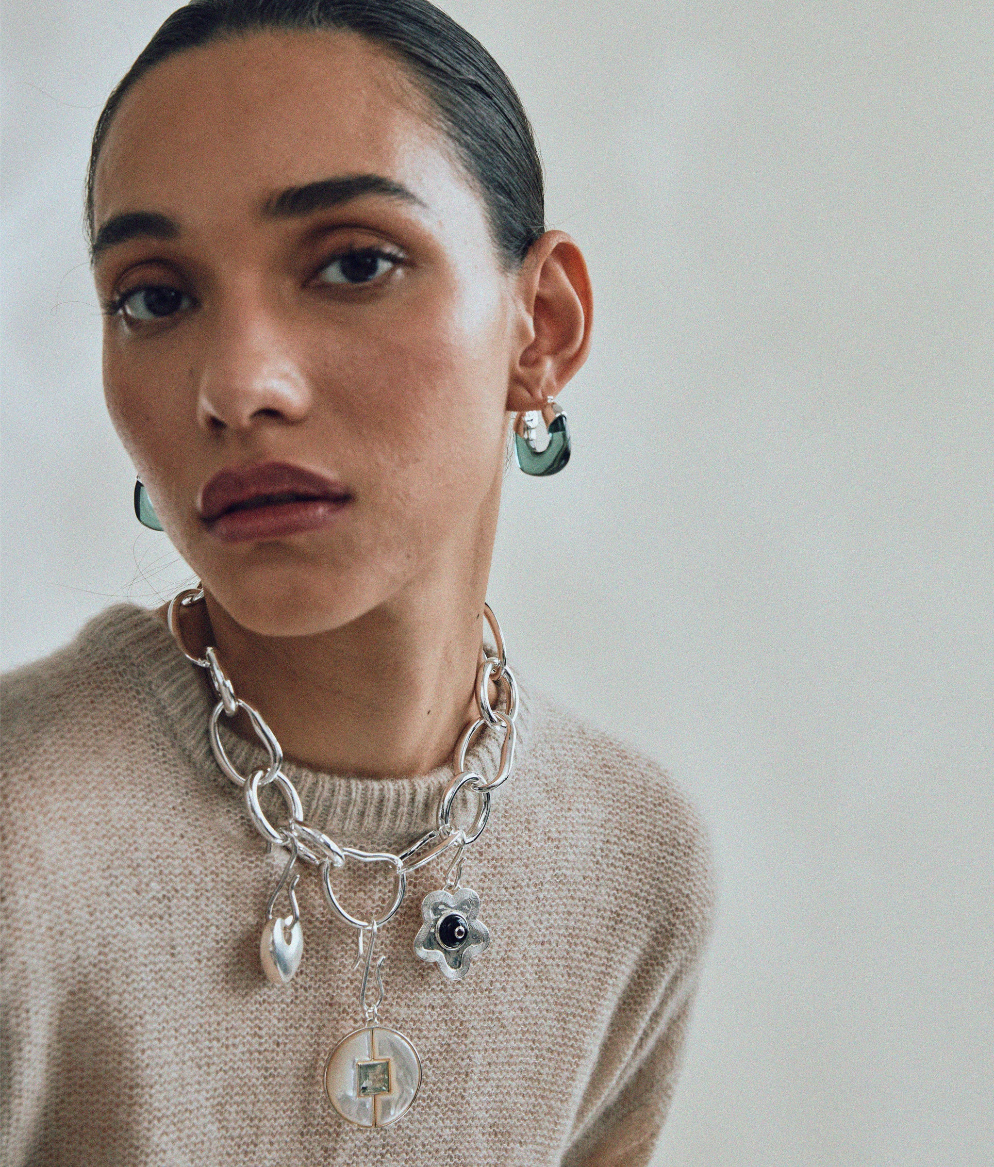 Model in tan sweater wear Porto Chain in Silver with attached silver charms, and Mini Organic Hoops in Forest & Silver.