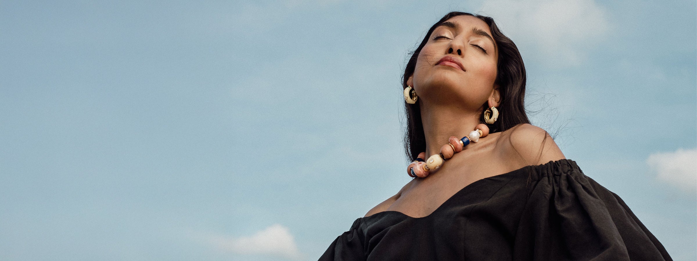 Model against blue sky wears beaded necklace and white hoop earrings with black top.