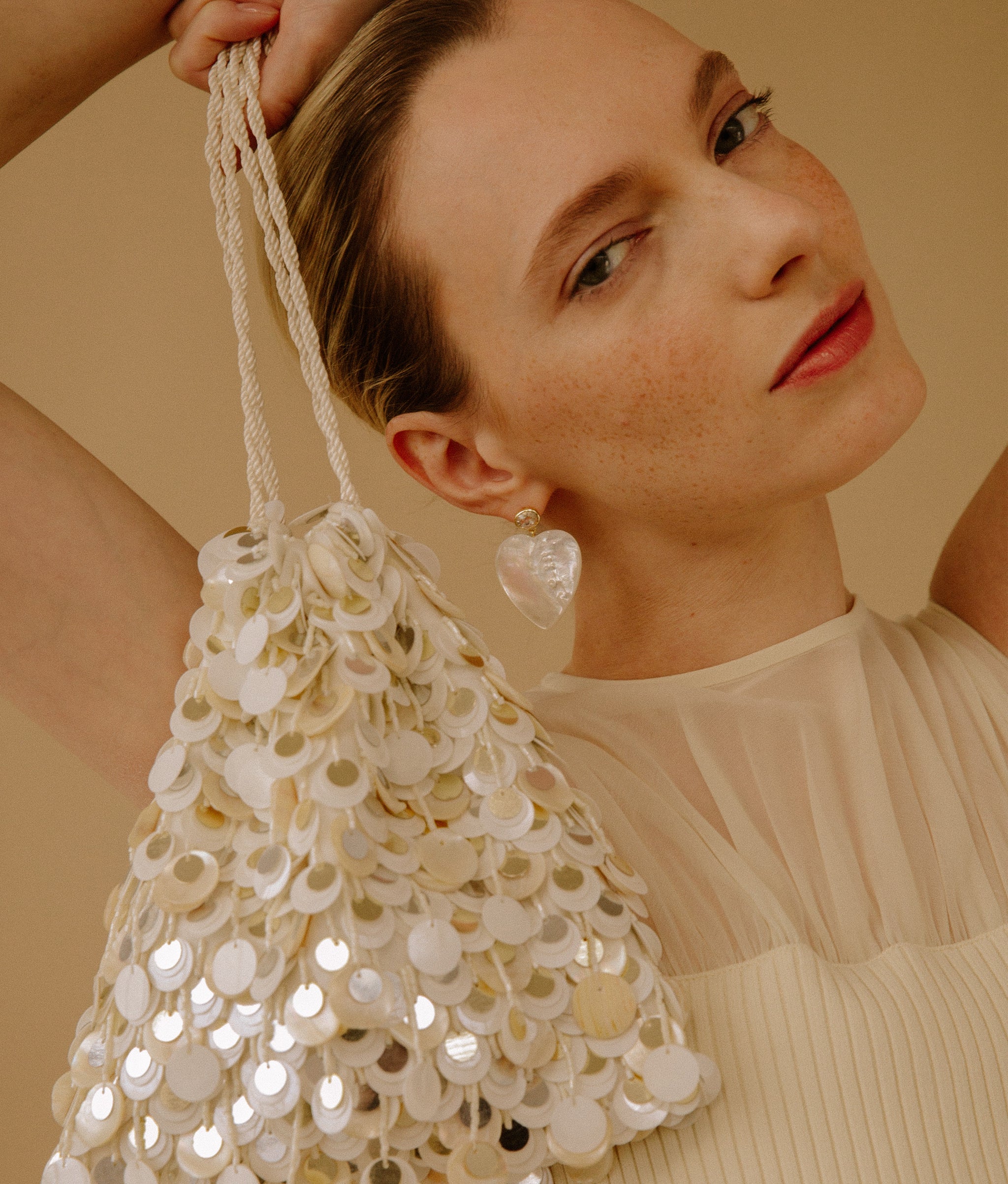 Model on a tan background carrying the Gala Bag in Pearl Oyster while wearing the Valentina Earrings.