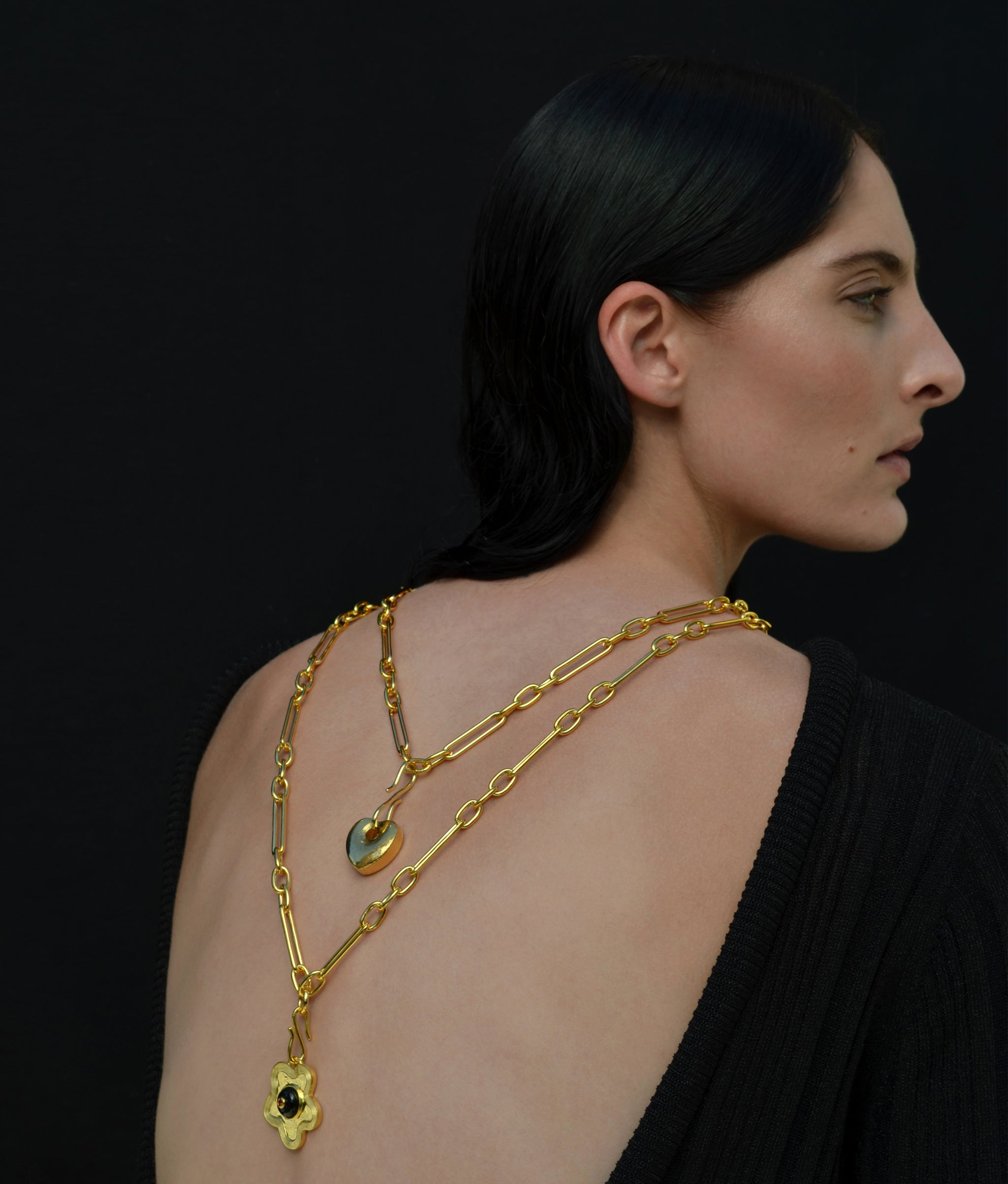 Model on black backdrop wears low-cut black dress and gold chains and charms turned backwards