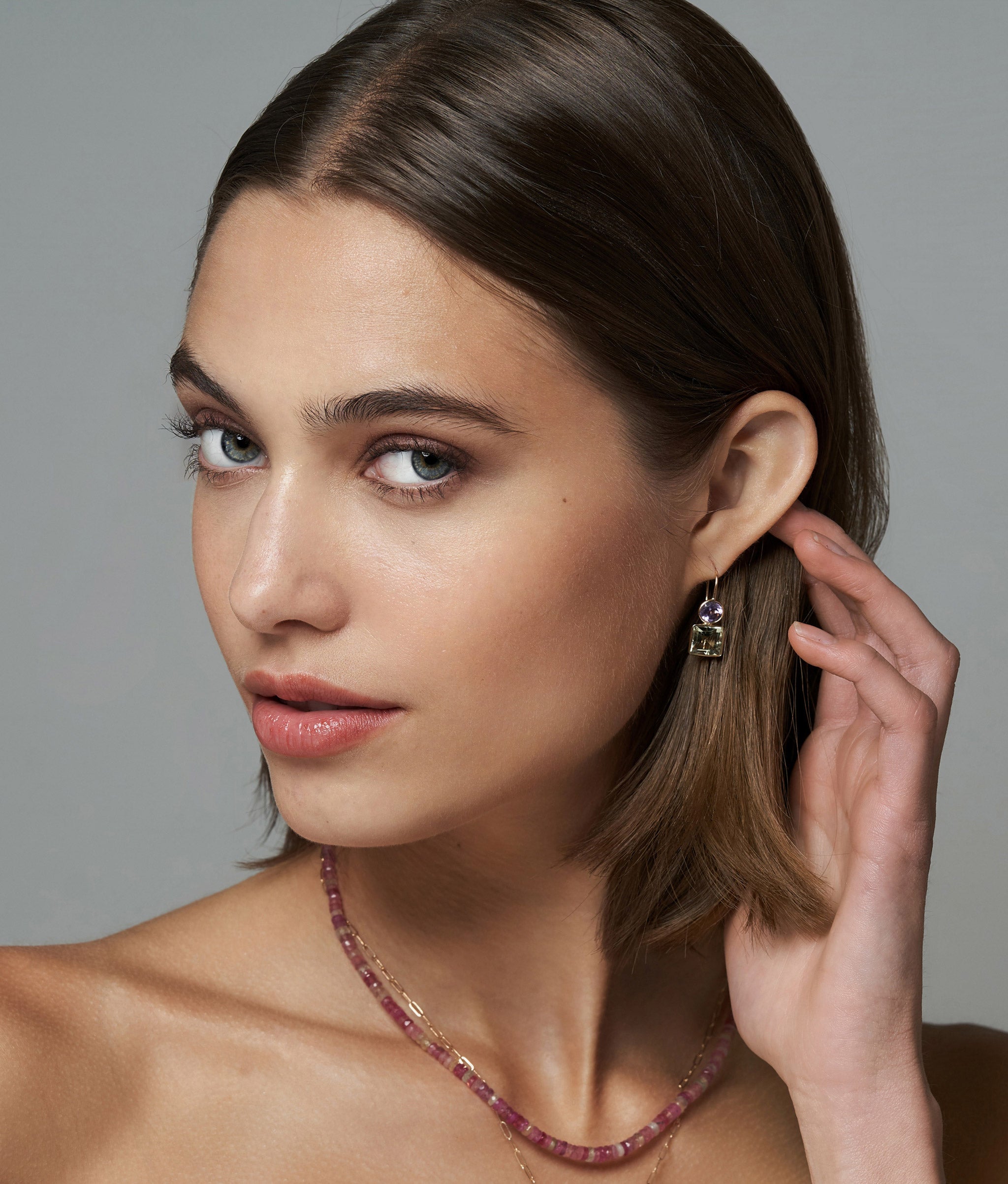 Model on grey backdrop wears 14k Pastille Earrings in Amethyst and Green Amethyst, and Pink Sapphire Necklace.