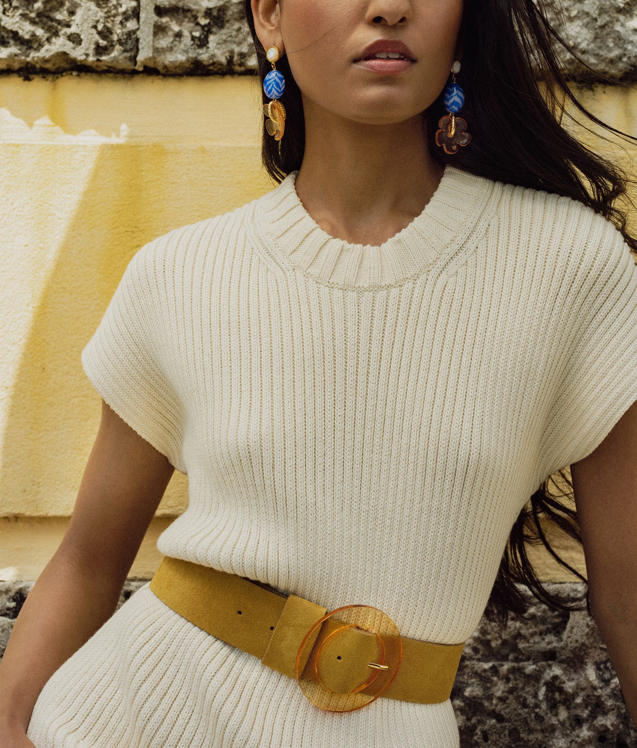 Model on yellow wall wears white ribbed dress, Mistflower Earrings in Blue Horizon, and Louise Belt in Ochre.