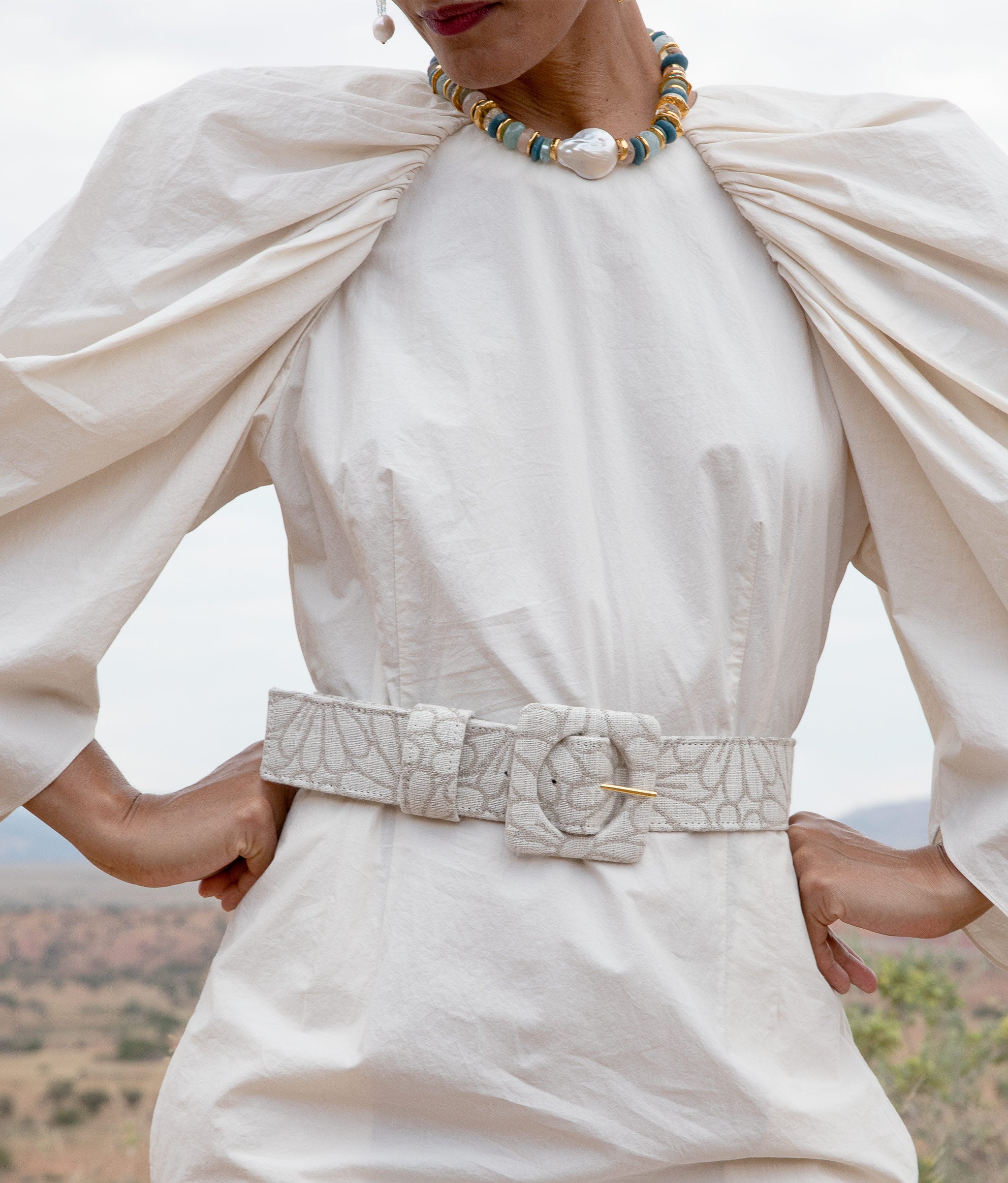Model with hands on hips wears white dress with dramatic puff sleeves, cinched by the Agnes Belt in Daisy Linen.