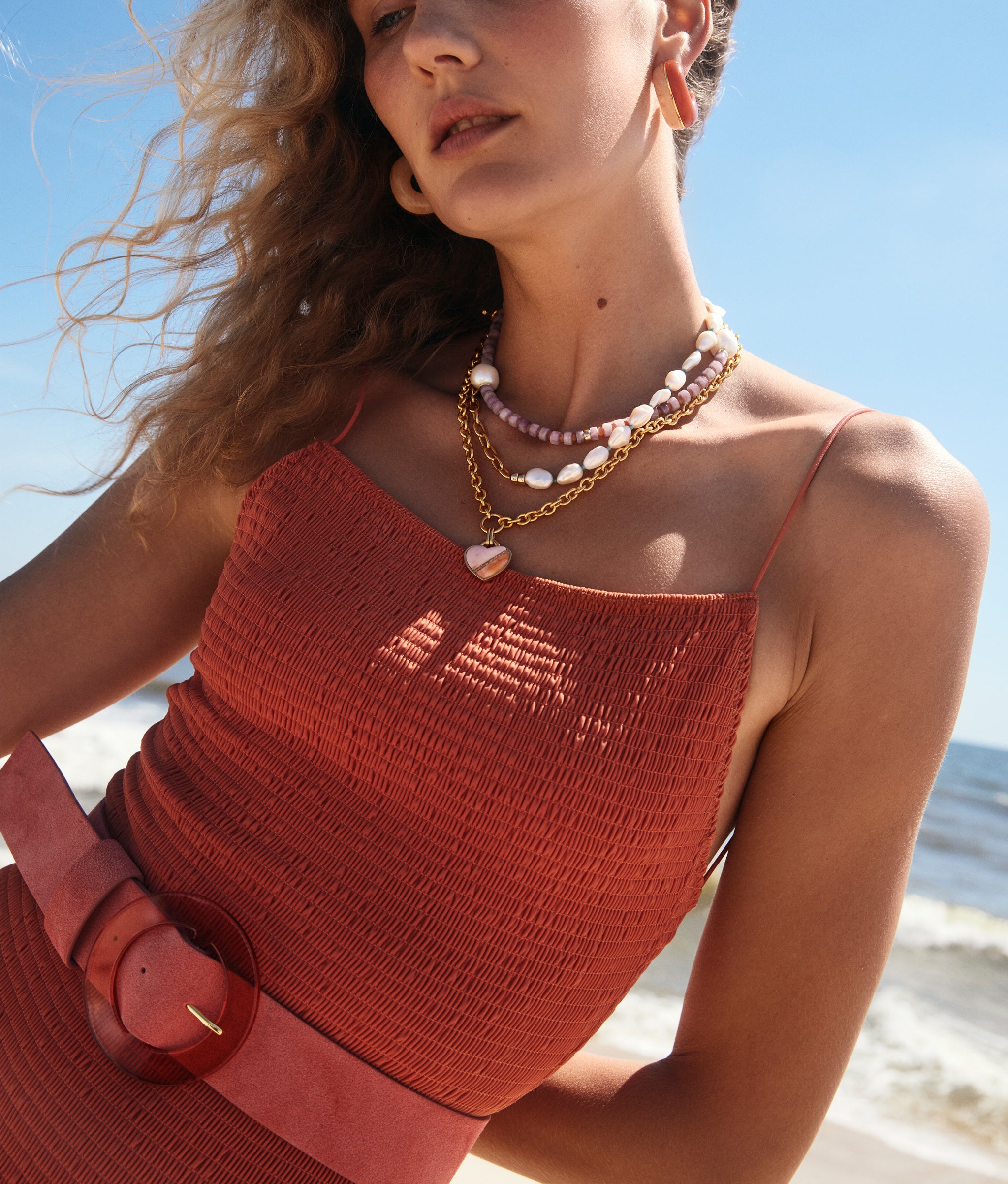 Model on beach wears coral-colored swimsuit with Louise Belt in Conch Suede and assorted jewelry.