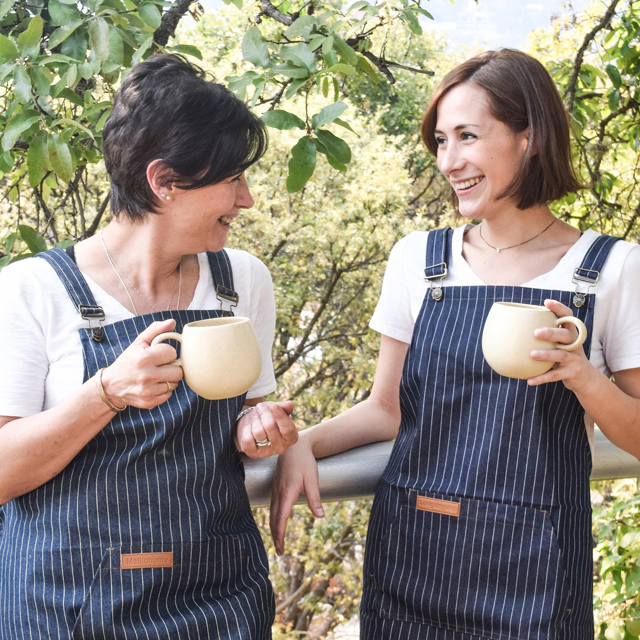 Two women in white t-shirts and navy striped aprons hold artisan mugs