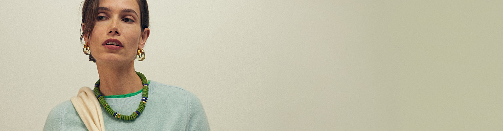 Model on tan backdrop wears blue sweater with Laguna Necklace in Pistachio and Organic Hoops