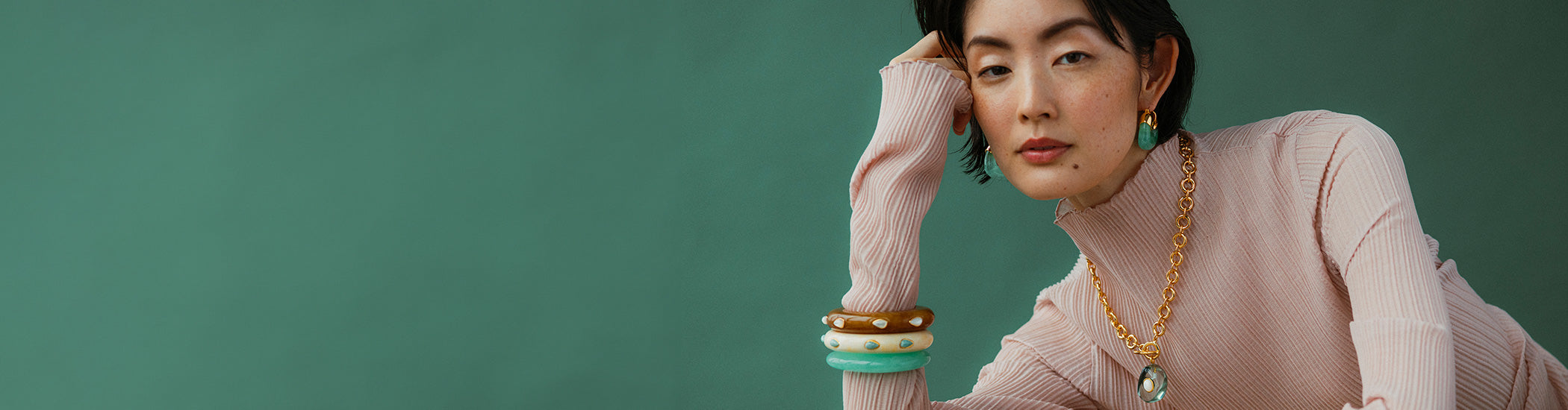 Model on green backdrop poses with hand on her head and stack of colorful resin bracelets