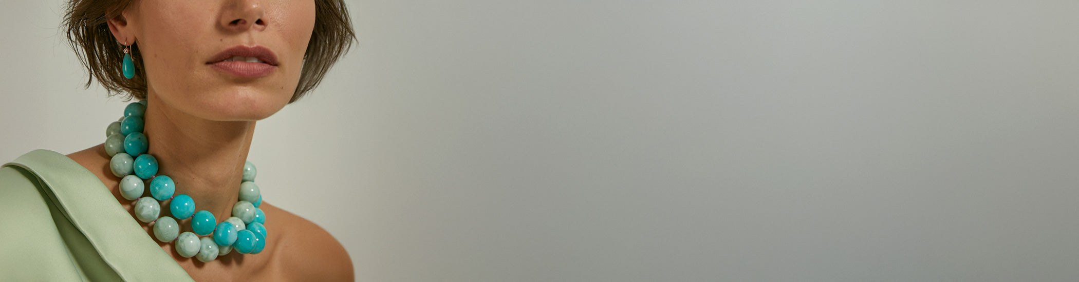 Model on grey backdrop wears one-shoulder green top with blue and green bead necklaces