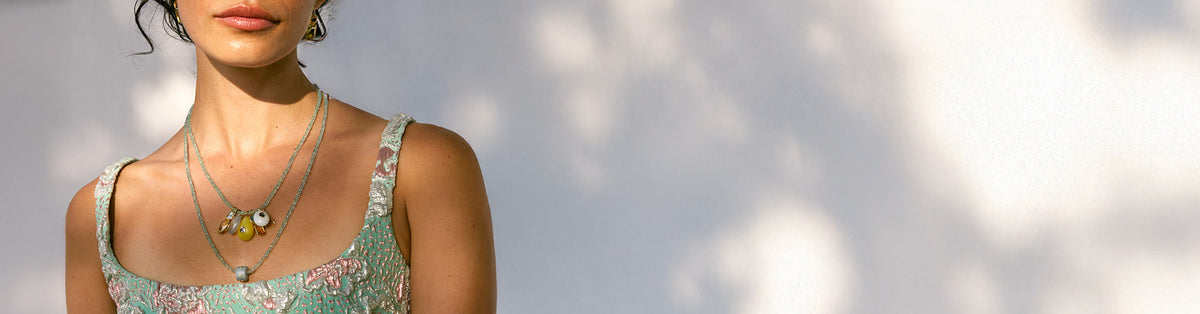 Model on grey sunlit backdrop wears aqua top with tiny emerald bead necklaces and charms