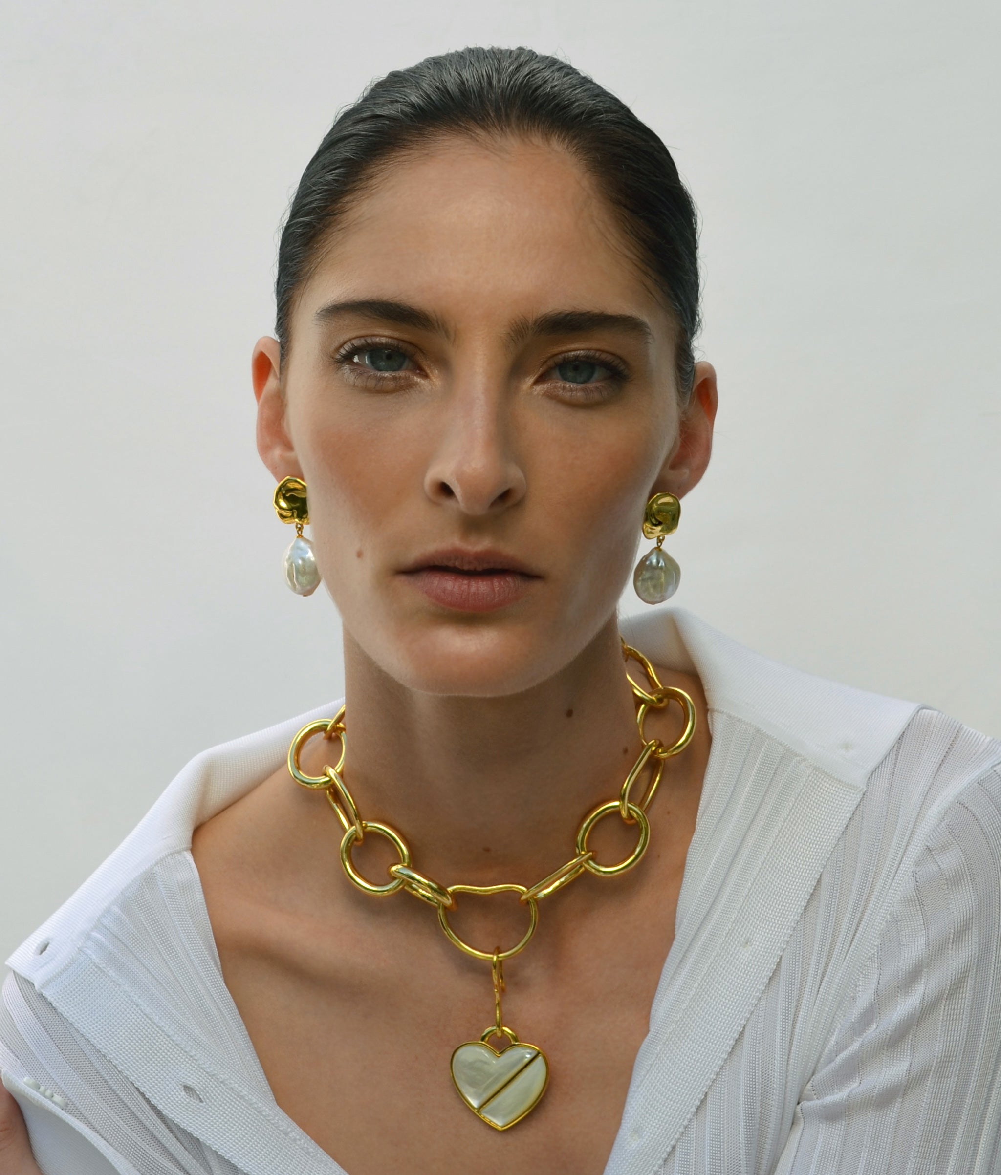 Model on grey backdrop wears white blouse with Porto Chain and heart charm and Coin Reflection Earrings