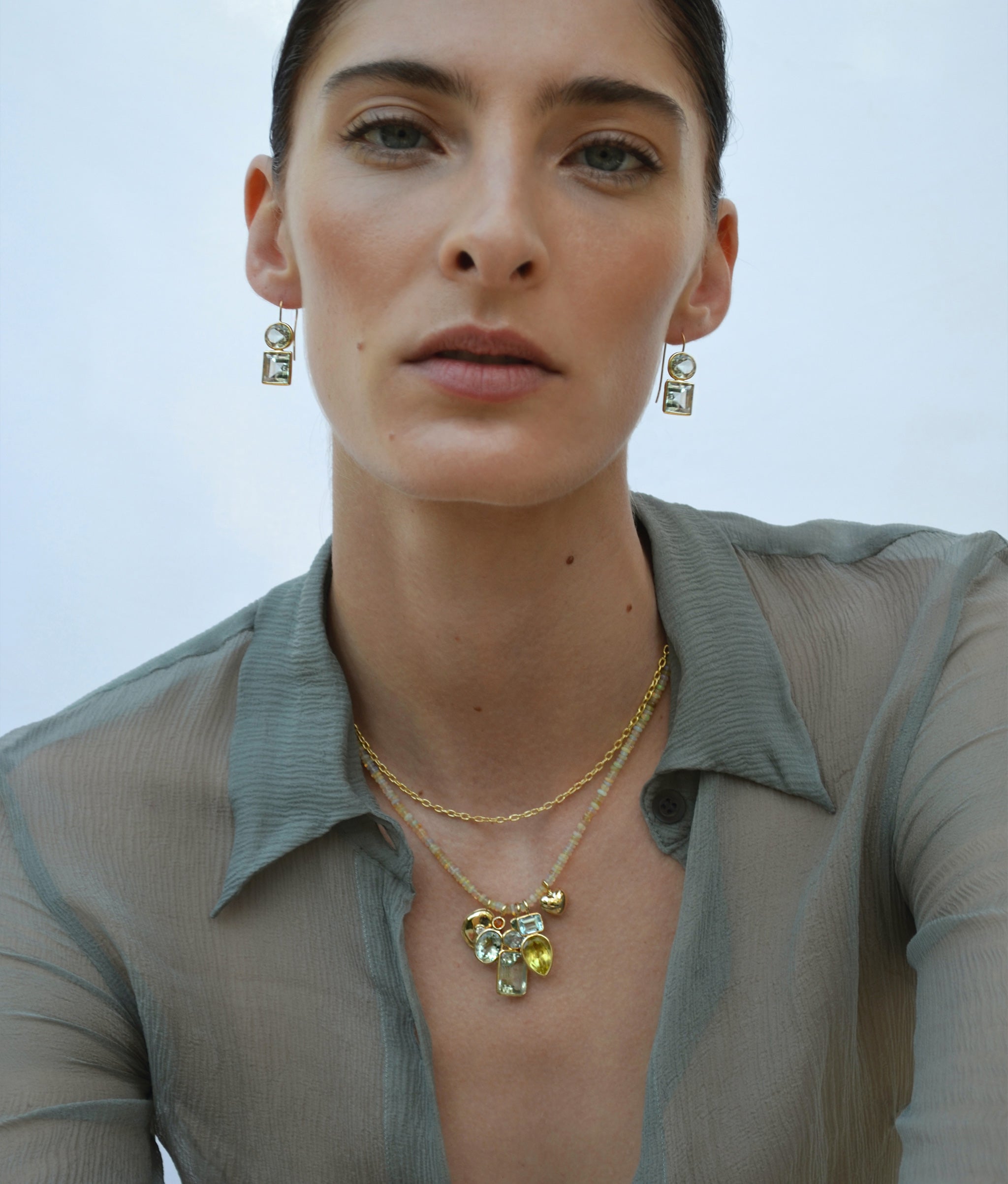 Model on blue backdrop wears gauzy top with beaded necklace and assorted semiprecious charms Model wears sage green transparent blouse with bead necklace and semiprecious charms