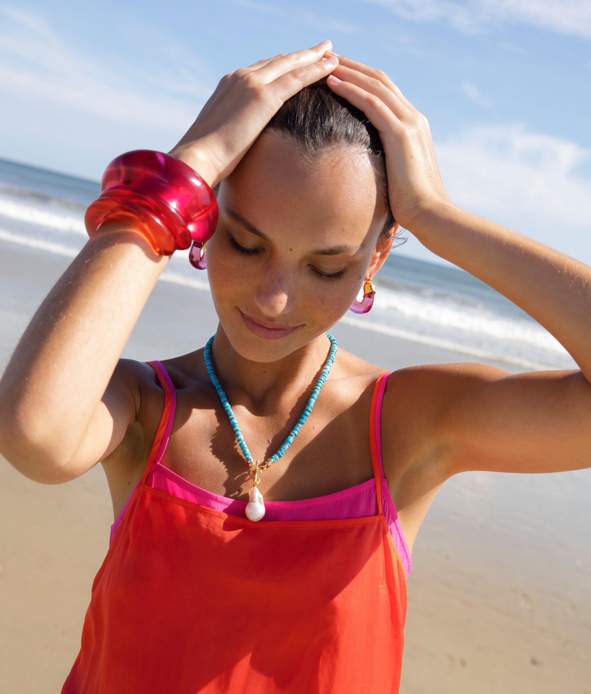 Model at the beach wearing Arc Cuff in Magenta paired with pink and orange cuffs, pink hoops, and a blue necklace.