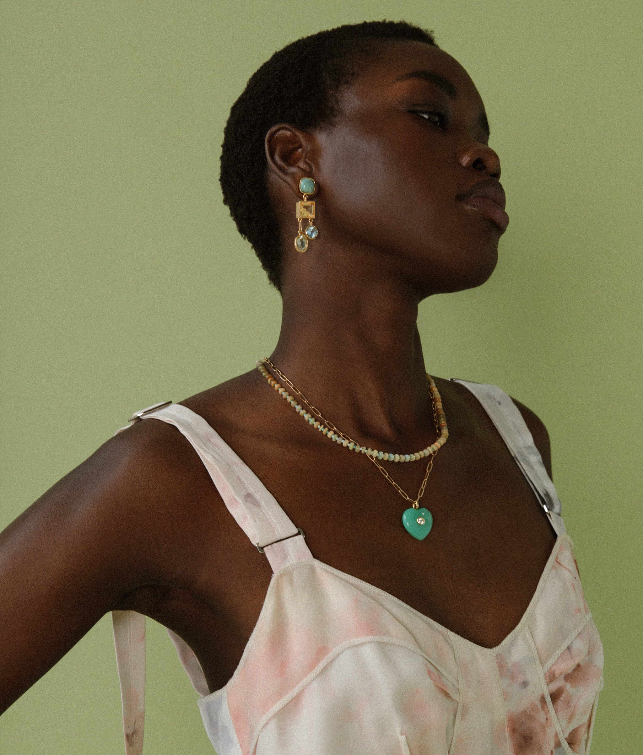 Model on green backdrop wears floral dress with Anemone Earrings and Lamai Heart Necklace.