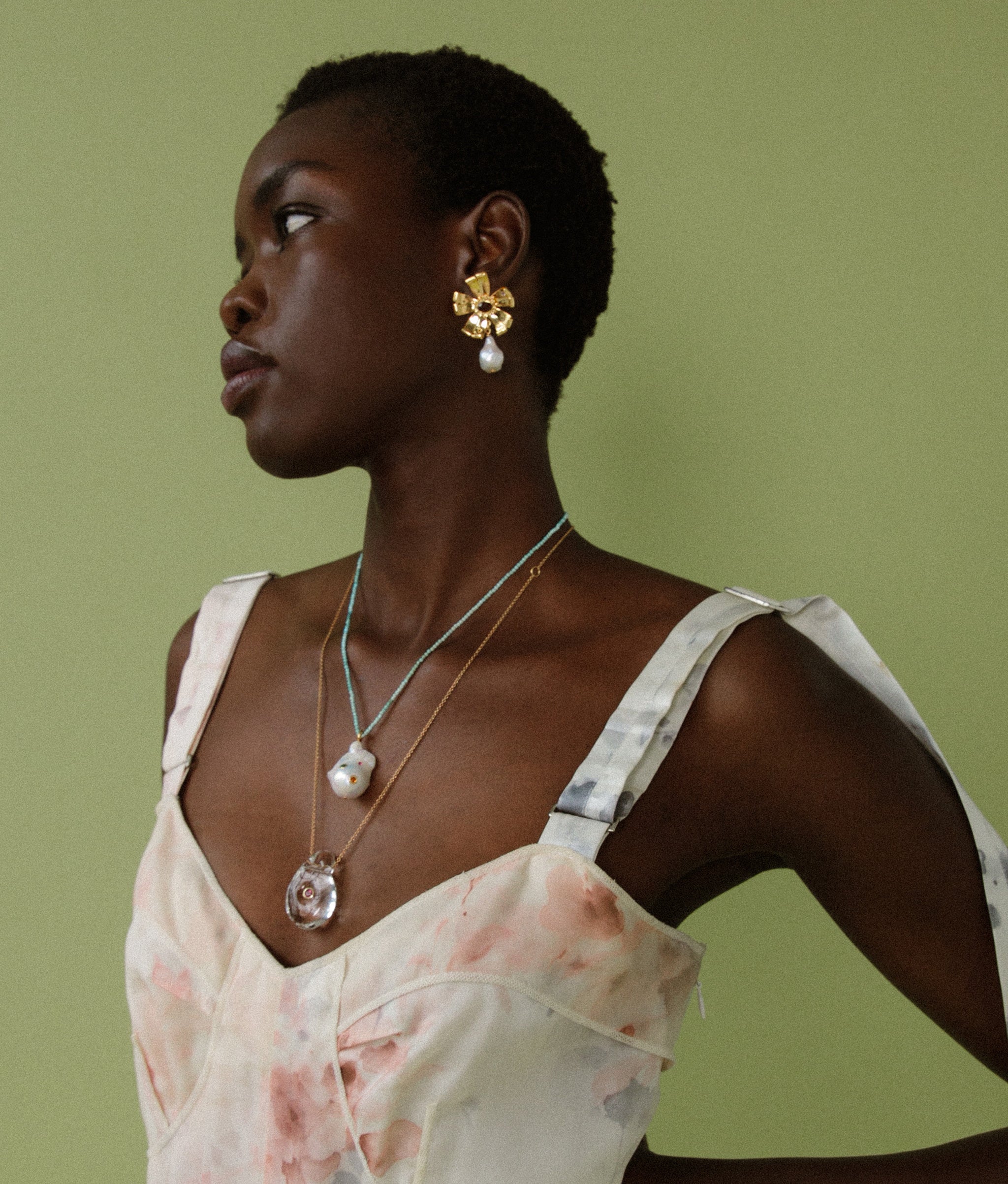 Model on green backdrop wears watercolor floral print dress with Lotus Earrings, Muse Pendant, and Skyfall Necklace.