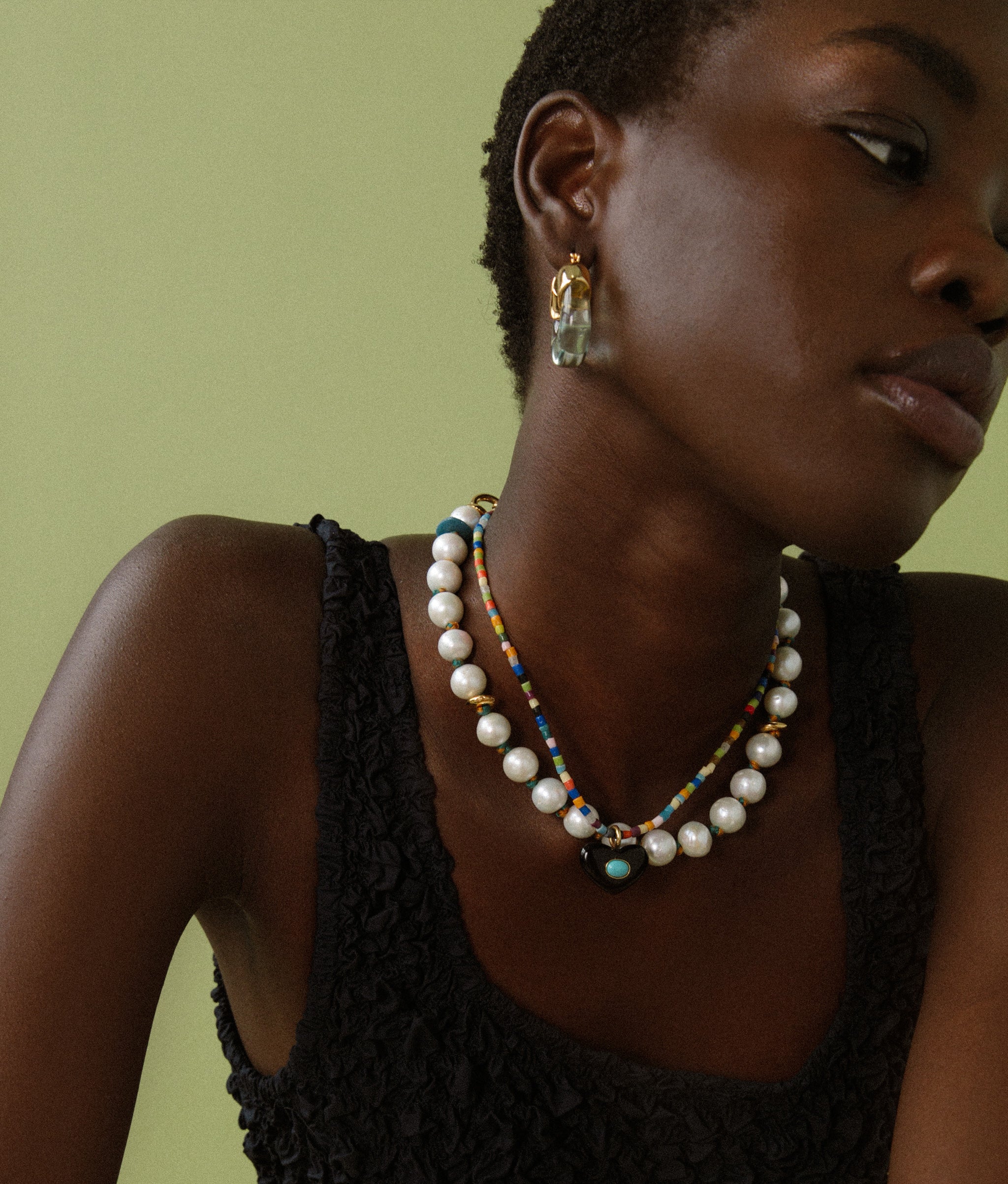 Model on green backdrop wears black tank with Martina Heart Necklace and Pacifica Pearl Collar. 