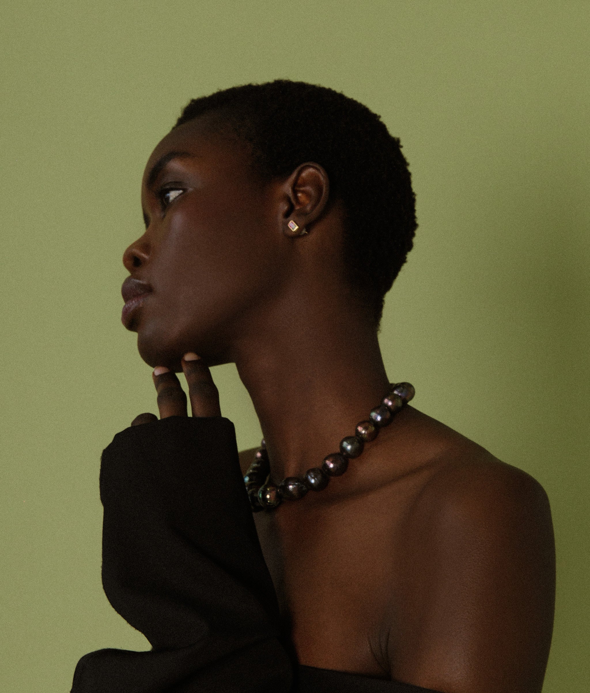 Model in profile on green backdrop with hand on chin wears Peacock Baroque Pearl & 14k Gold Necklace