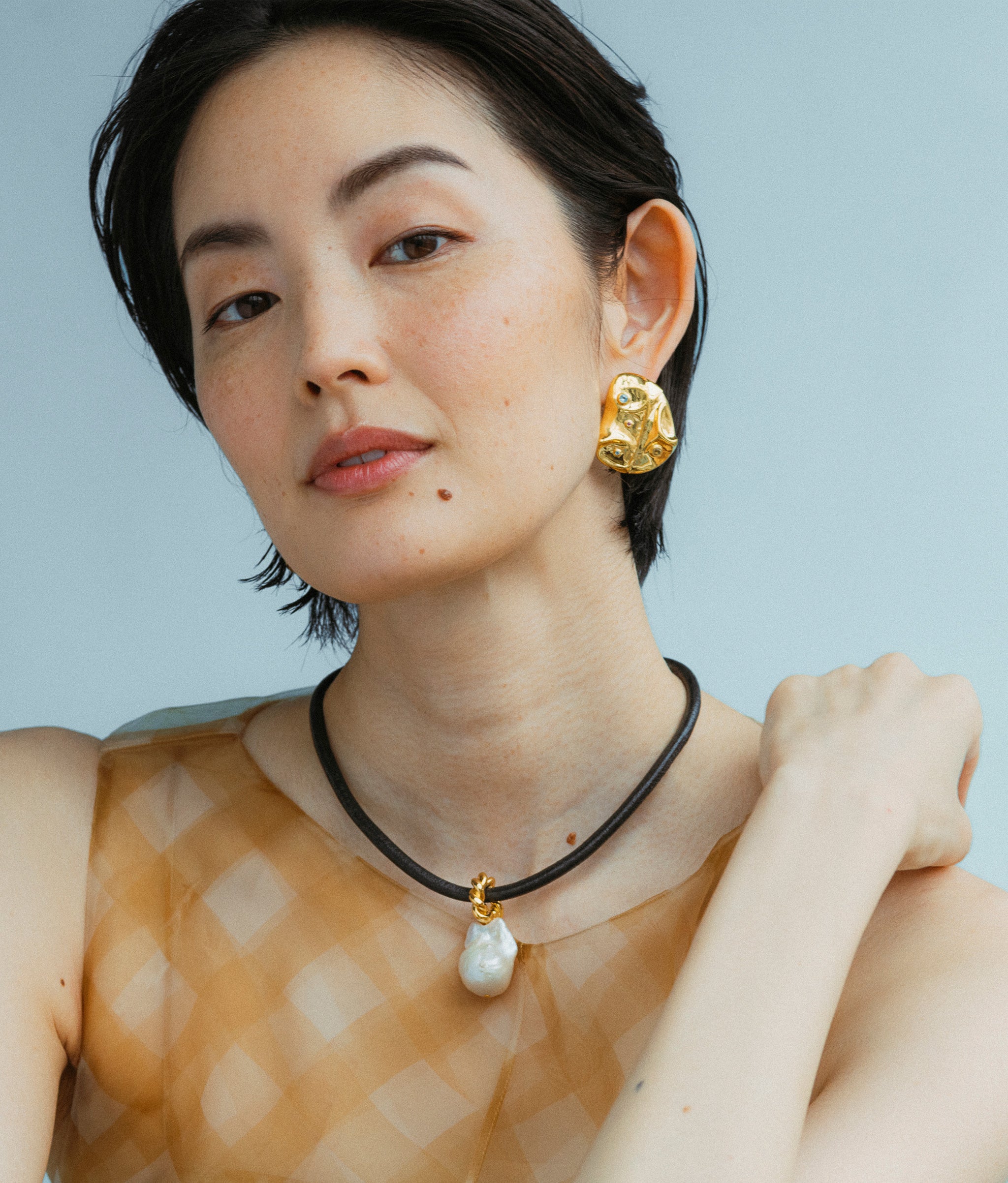 Model wears Esme Collar and Mazu Studs earrings in a blue backdrop. 