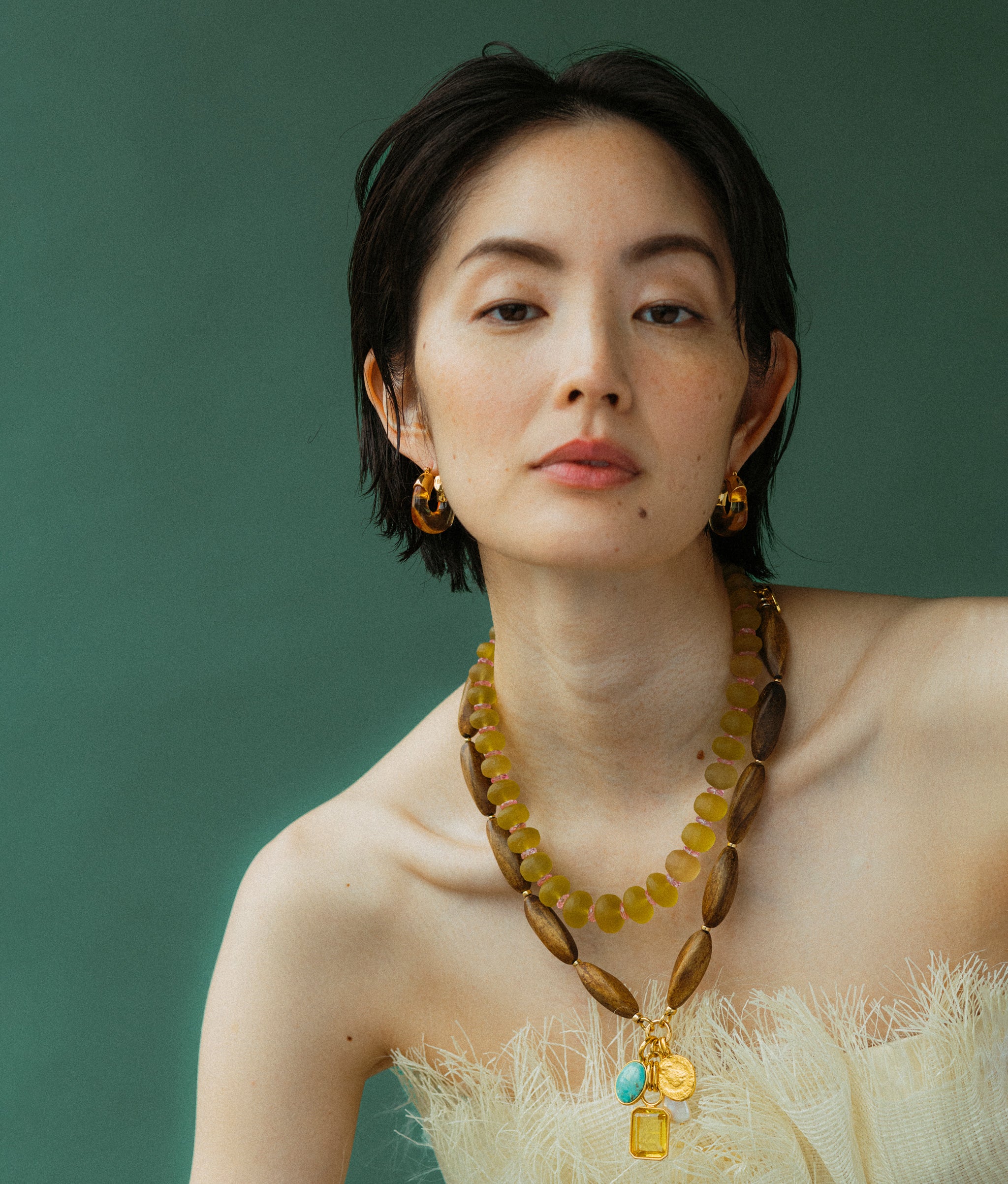 Model on a green backdrop wears Mini Organic Hoops In Honey featured with Marilla Necklace and Canary Cliffs Necklace.