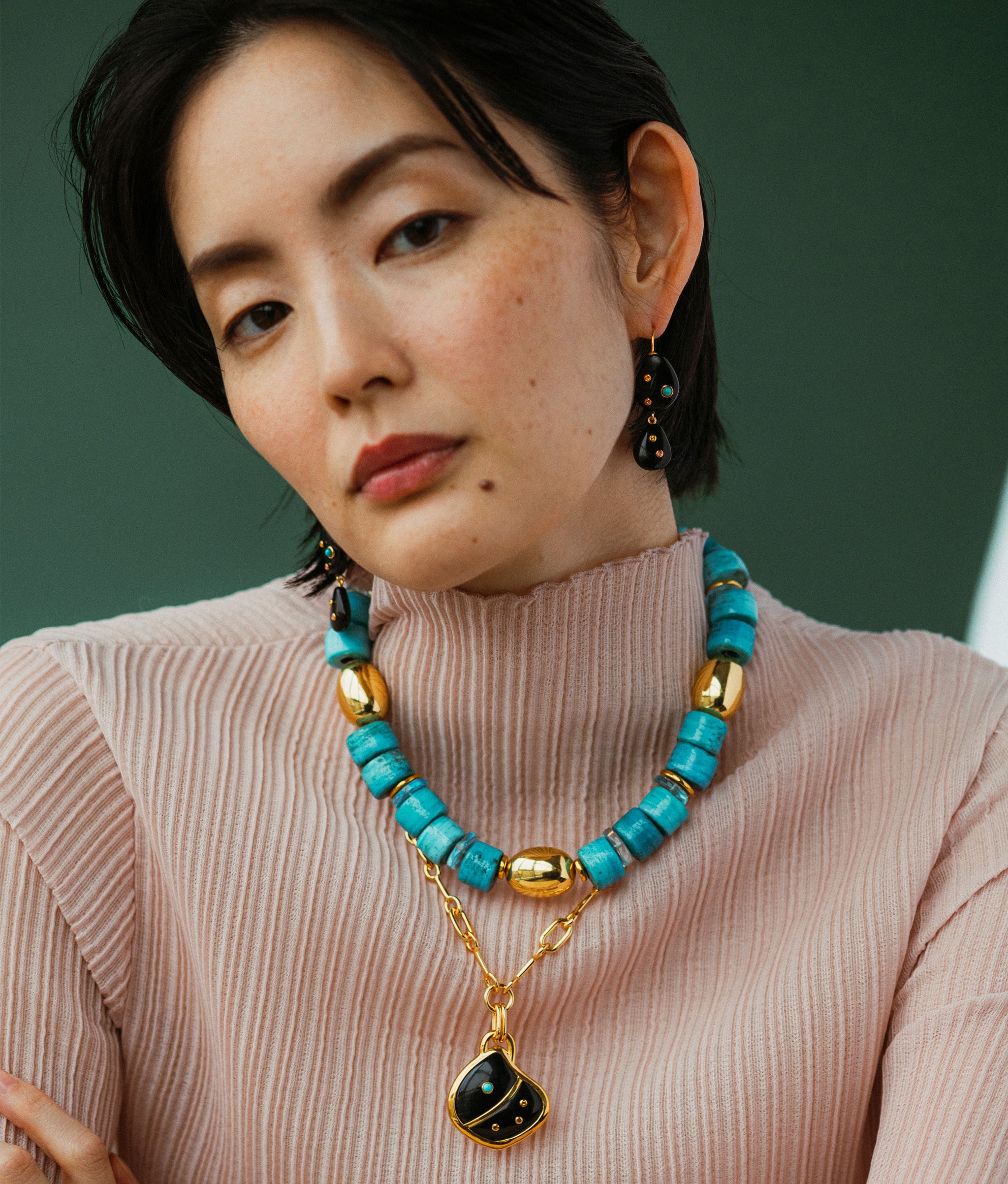 Model wears Stacked Stones Earrings in Black Agate paired with necklaces on a green backdrop. 