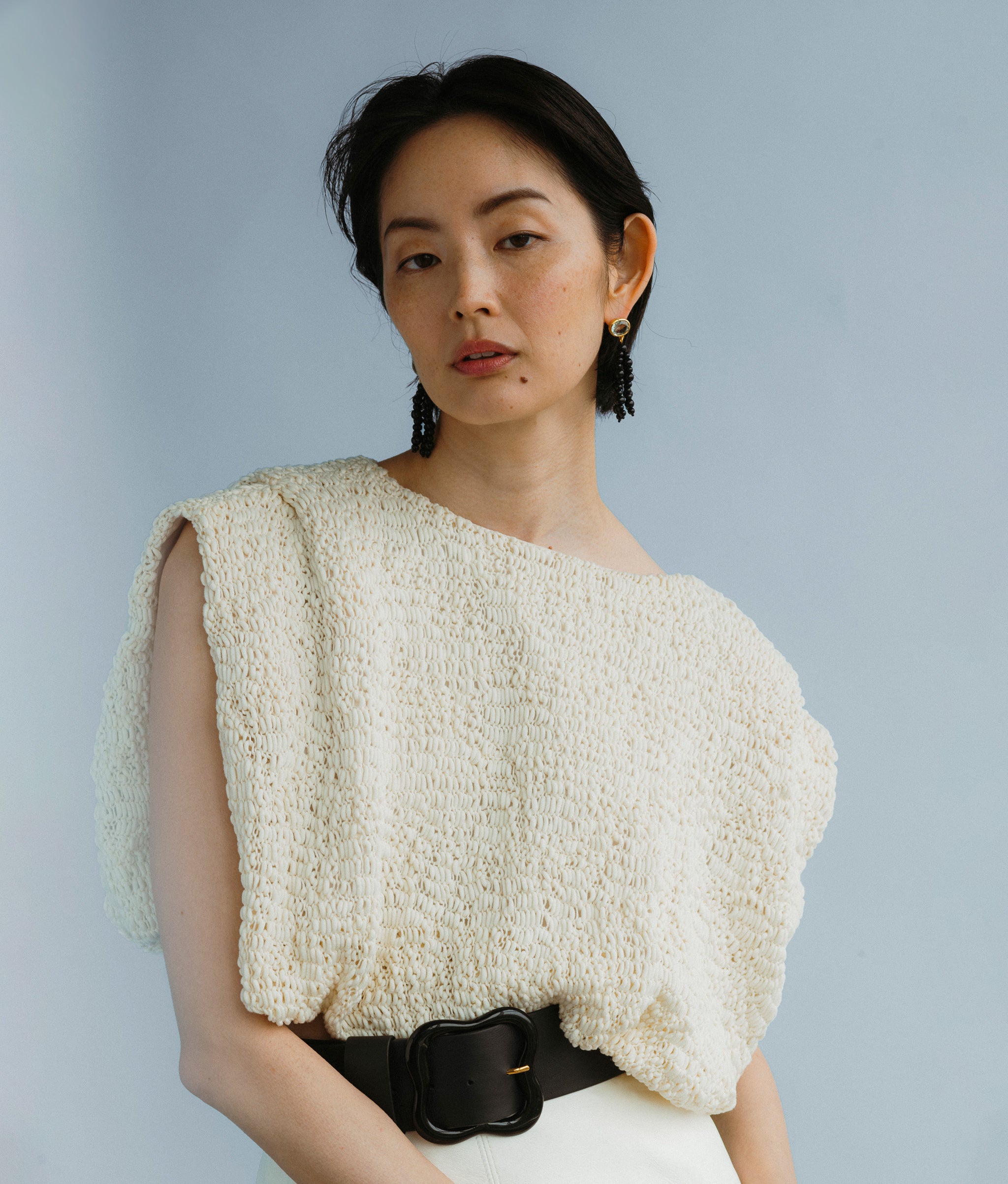 Model wears a white top along with Midnight Masquerade Earrings and Florence Belt In Jet black on a blue backdrop.