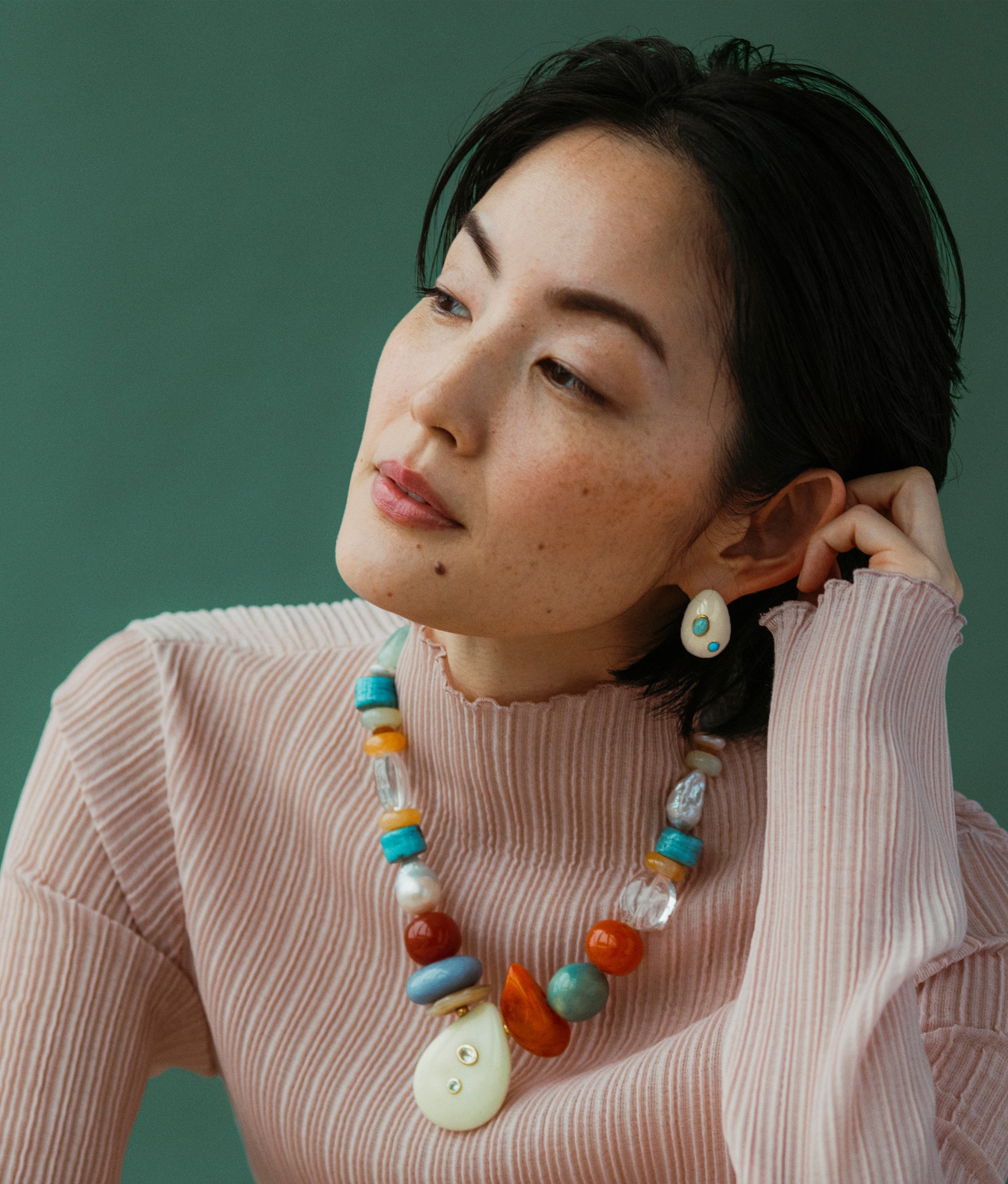 Model wears Artifact Necklace and Mini Arp Earrings in Studded Cream in a green background.