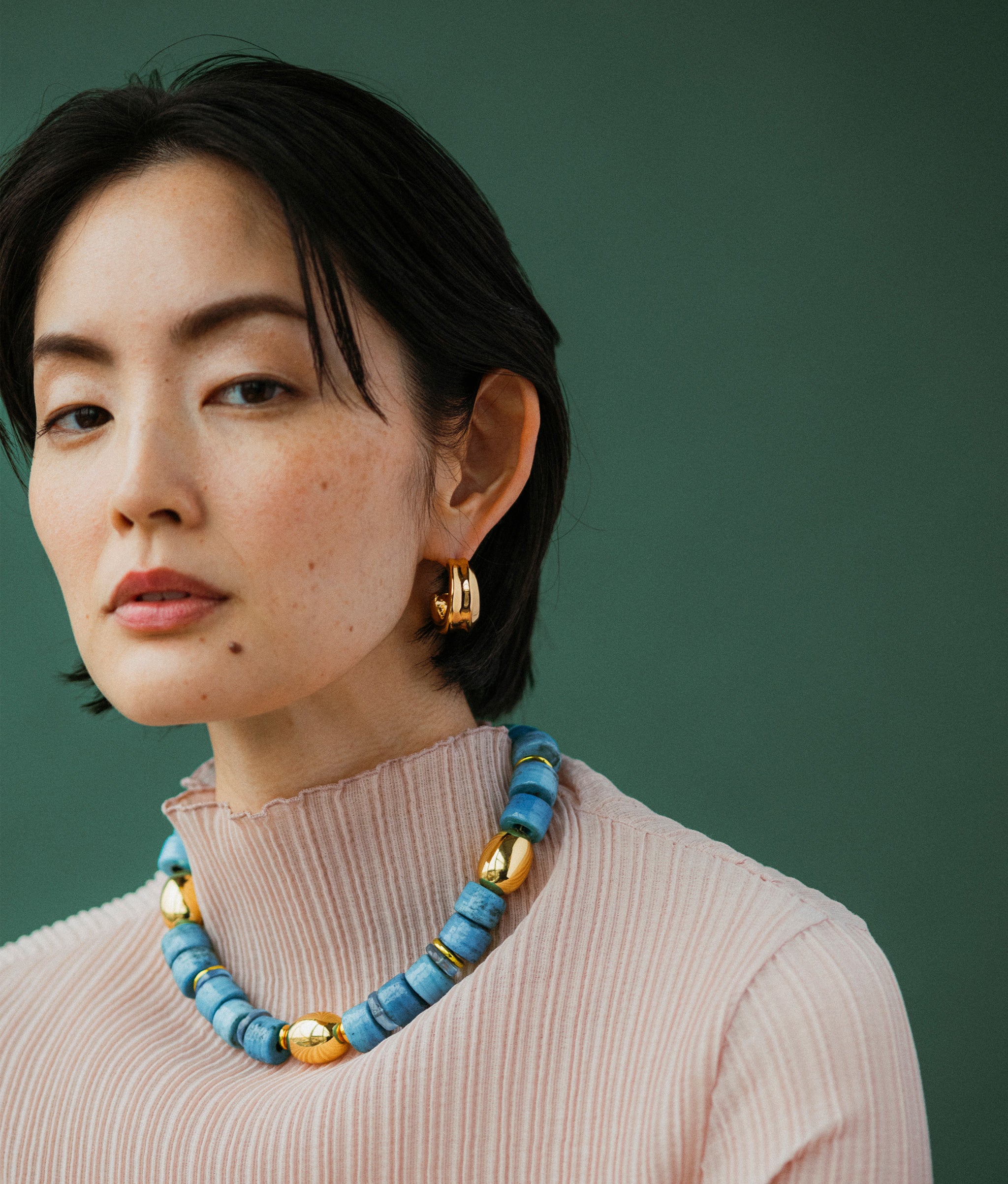 Model wears Varuna collar and Onda Hoops In Gold on a green backdrop.