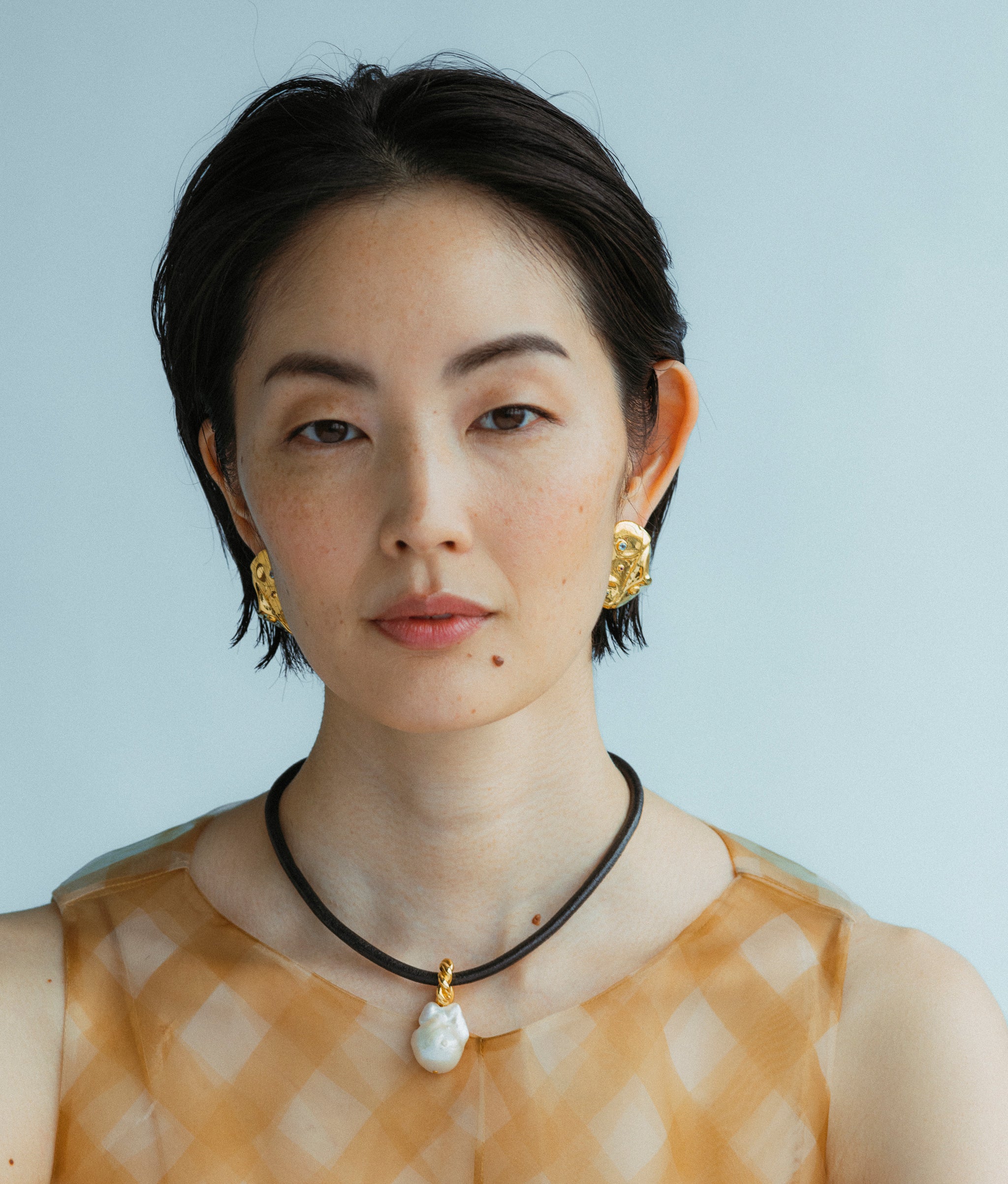 Model wears Esme Collar and Mazu Studs earrings in a blue backdrop. 