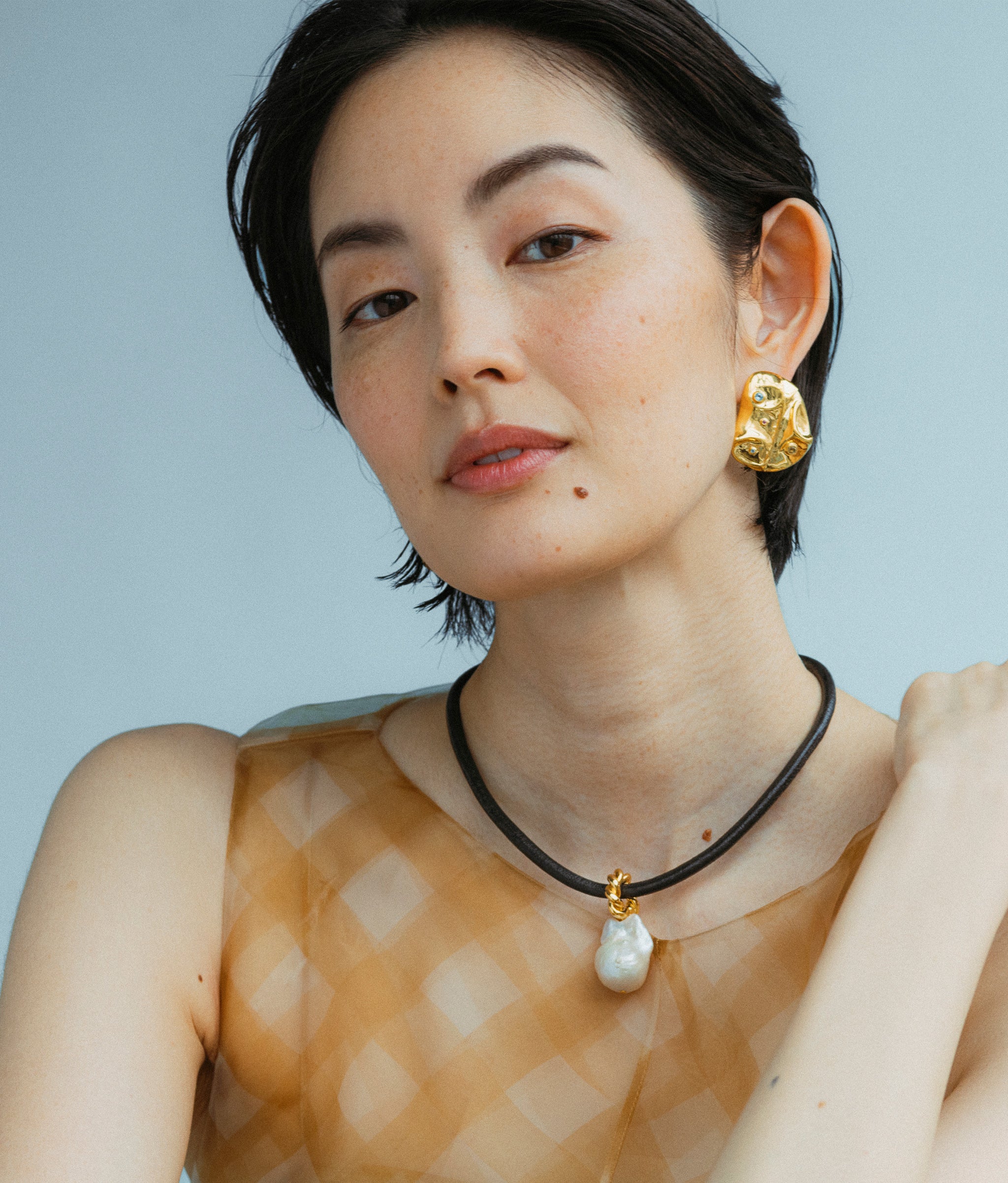 Model wears Esme Collar and Mazu Studs earrings in a blue backdrop. 