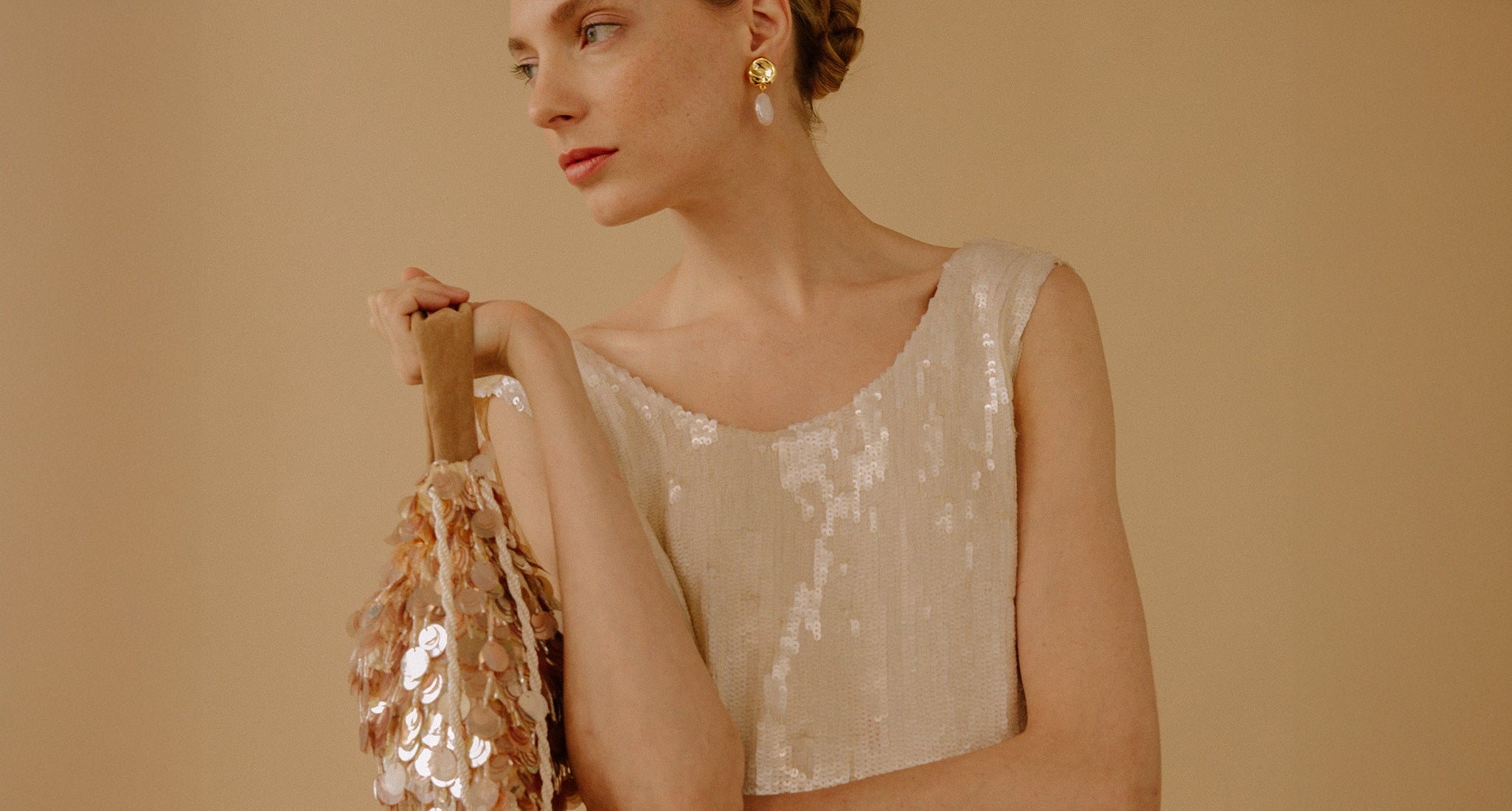 Model on tan backdrop wears white sequin top with Gala Bag and Coin Reflection Earrings